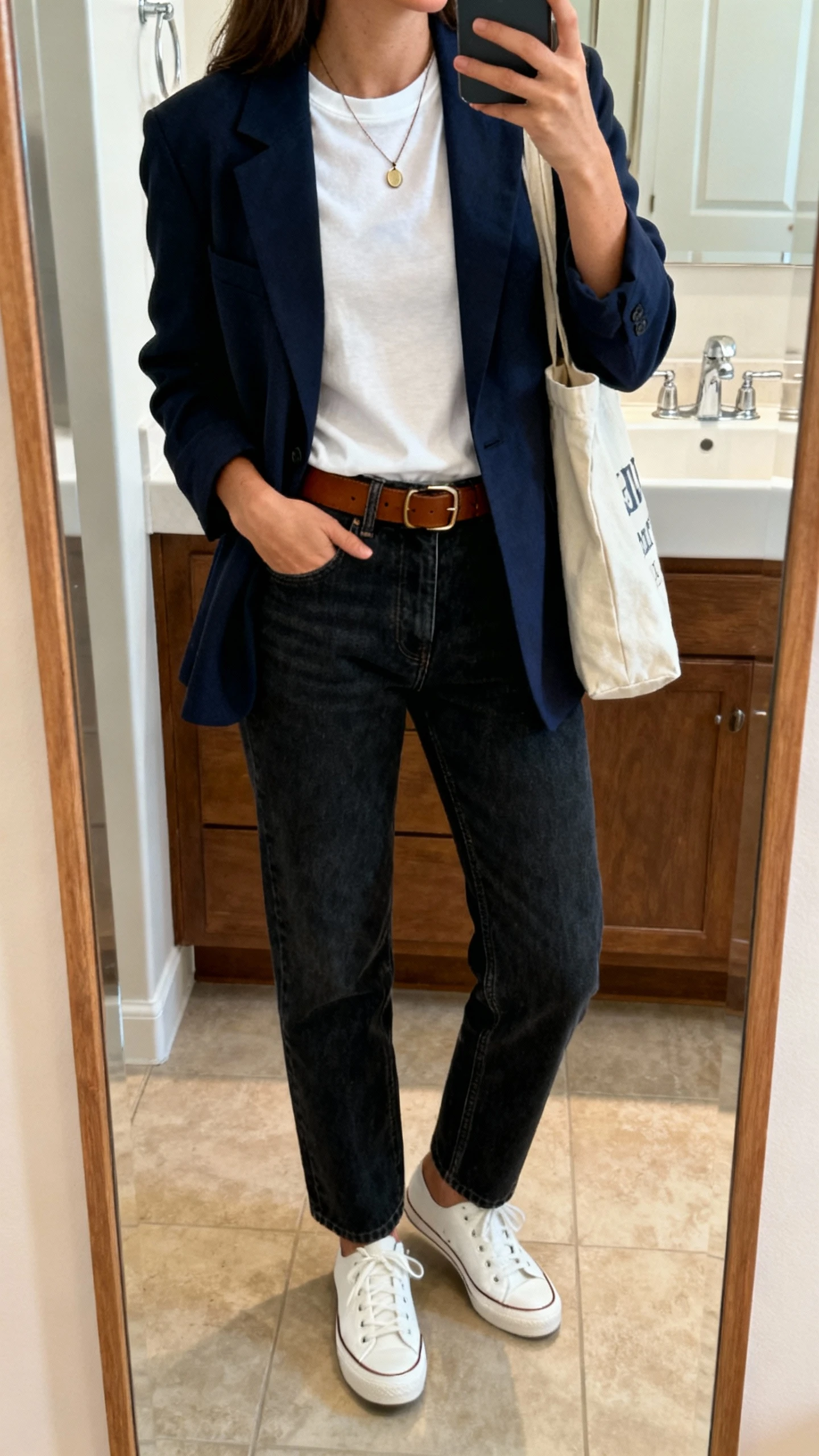 iPhone mirror selfie of a woman wearing a dark-wash straight-leg jean, relaxed navy blazer, white crewneck tee, brown leather belt, white leather sneakers, simple pendant necklace and tote bag, face not visible, bathroom mirror, warm indoor lighting, natural iPhone photo quality.