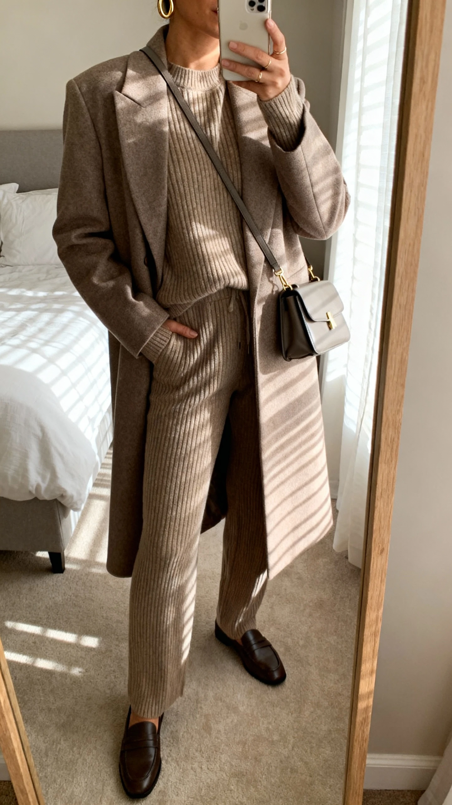 iPhone mirror selfie of a woman wearing a matching luxe knit set (ribbed sweater and knit pants) with a long tailored coat, minimalist gold hoops, sleek crossbody bag, and loafers, face not visible, cropped at neck, bedroom mirror with morning window light, natural iPhone photo quality.