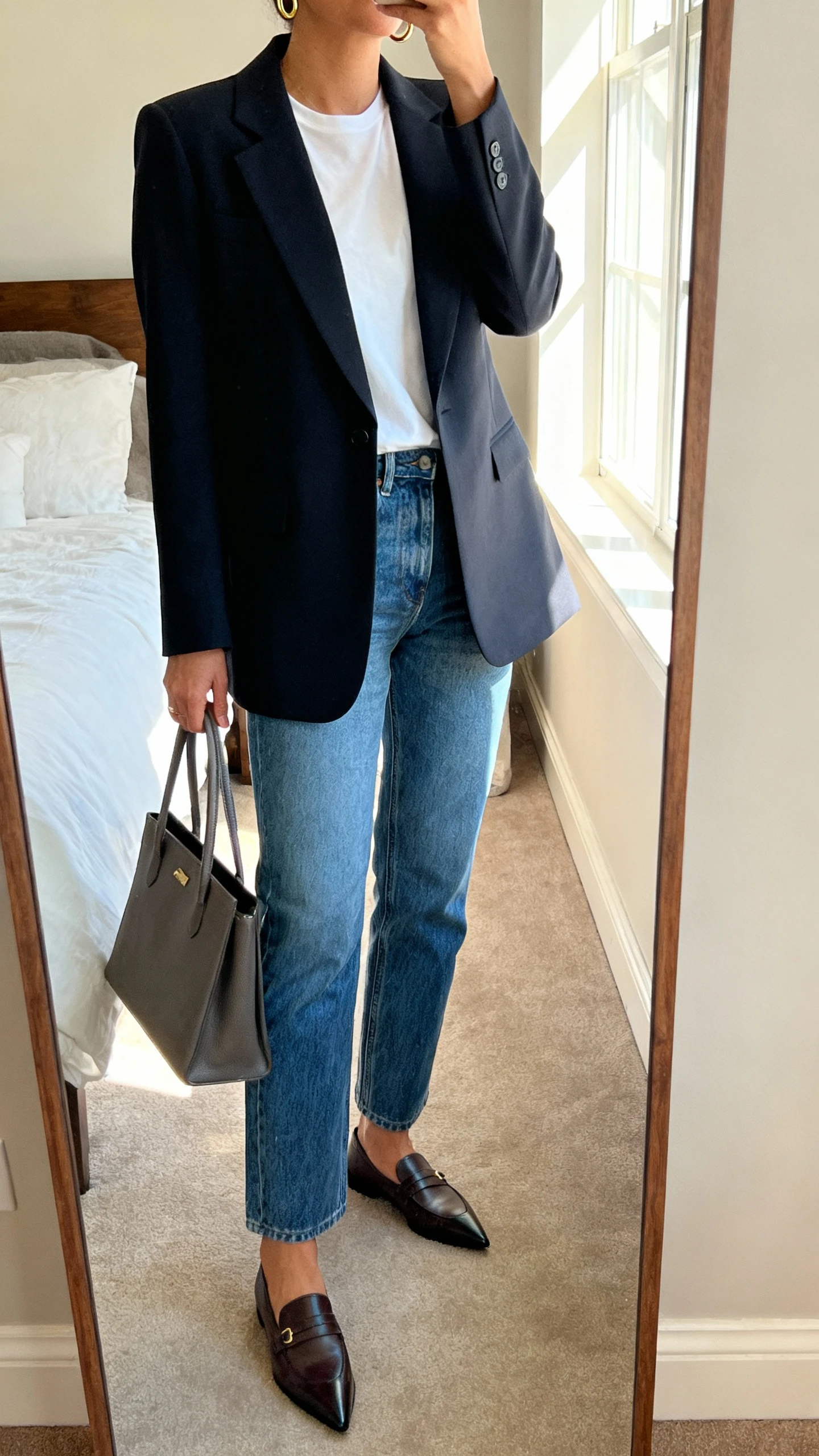 iPhone mirror selfie of a woman wearing a sharp power blazer over a white tee, straight-leg blue denim, pointed-toe loafers, a structured tote, and minimal gold hoops, face not visible, cropped at neck, bedroom mirror with morning window light, natural iPhone photo quality.