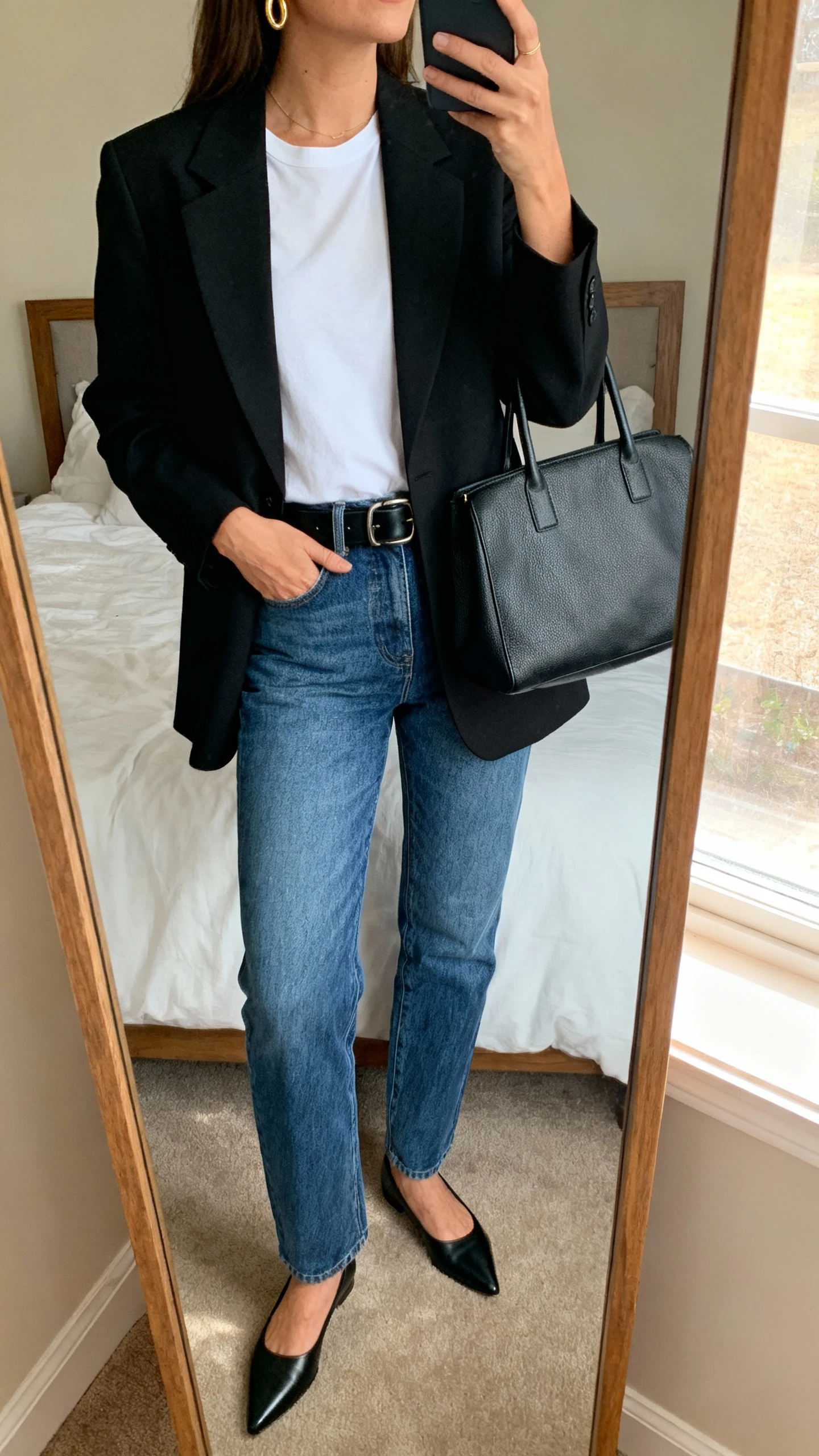 iPhone mirror selfie of a woman wearing a tailored black blazer over a white tee, straight-leg blue jeans, black leather belt, pointed loafers, dainty gold hoops, structured tote, face not visible, cropped at neck, bedroom mirror, natural window light, natural iPhone photo quality.