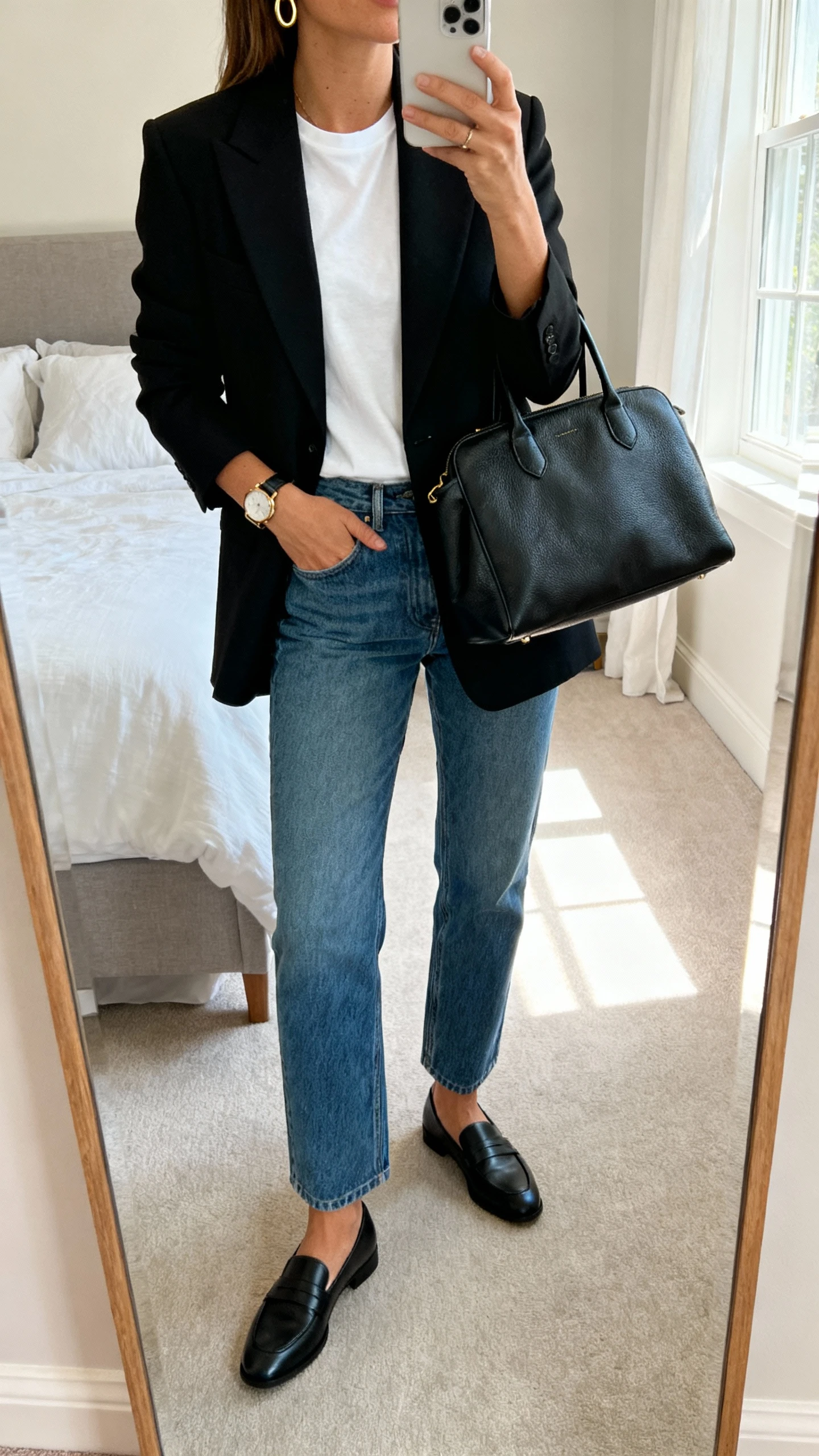 iPhone mirror selfie of a woman wearing a tailored black blazer over a white tee, straight-leg blue denim, black loafers, structured leather tote, minimal gold hoops and watch; bedroom mirror with morning window light, face not visible, cropped at neck, natural iPhone photo quality.