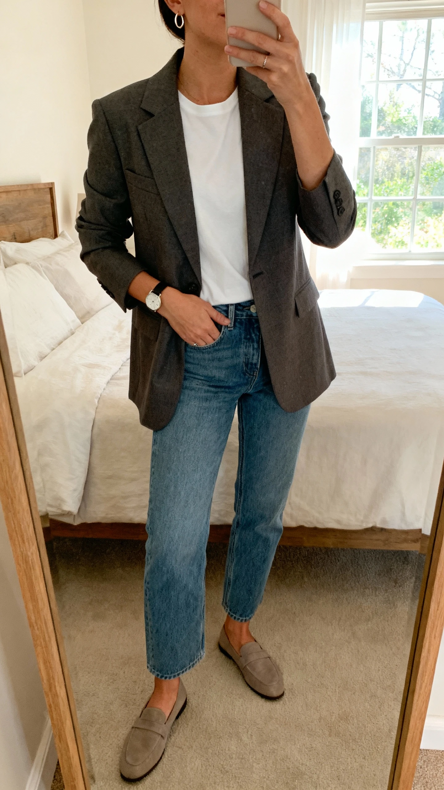 iPhone mirror selfie of a woman wearing a tailored blazer over a white tee with straight‑leg denim and minimalist loafers, simple watch and small hoop earrings, face not visible, cropped at neck, bedroom mirror with natural window light, natural iPhone photo quality.