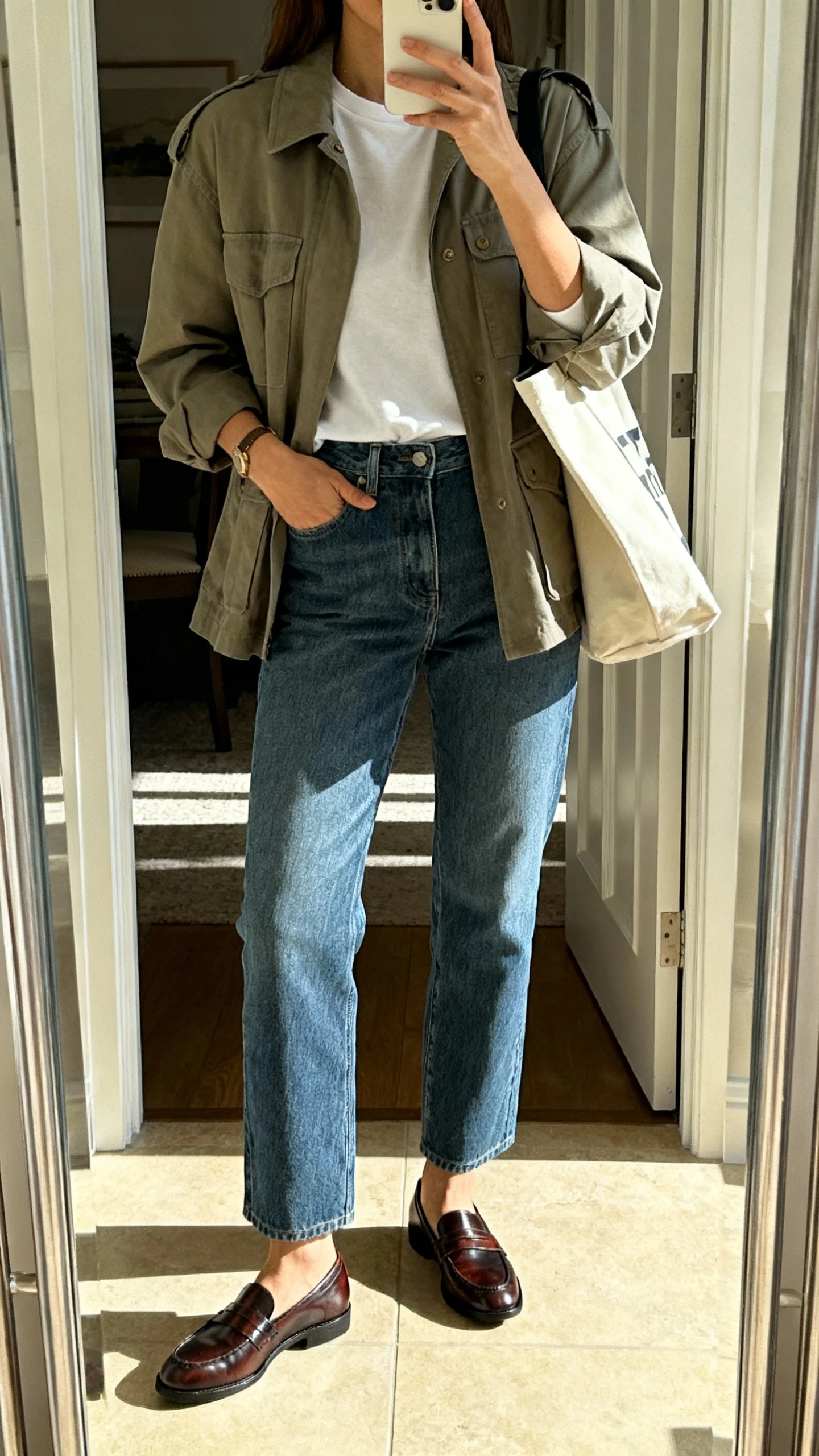 iPhone mirror selfie of a woman wearing a utility jacket over a crewneck tee, relaxed straight jeans, polished slip-on loafers, and a tote, face not visible, entryway mirror with afternoon natural light, natural iPhone photo quality.