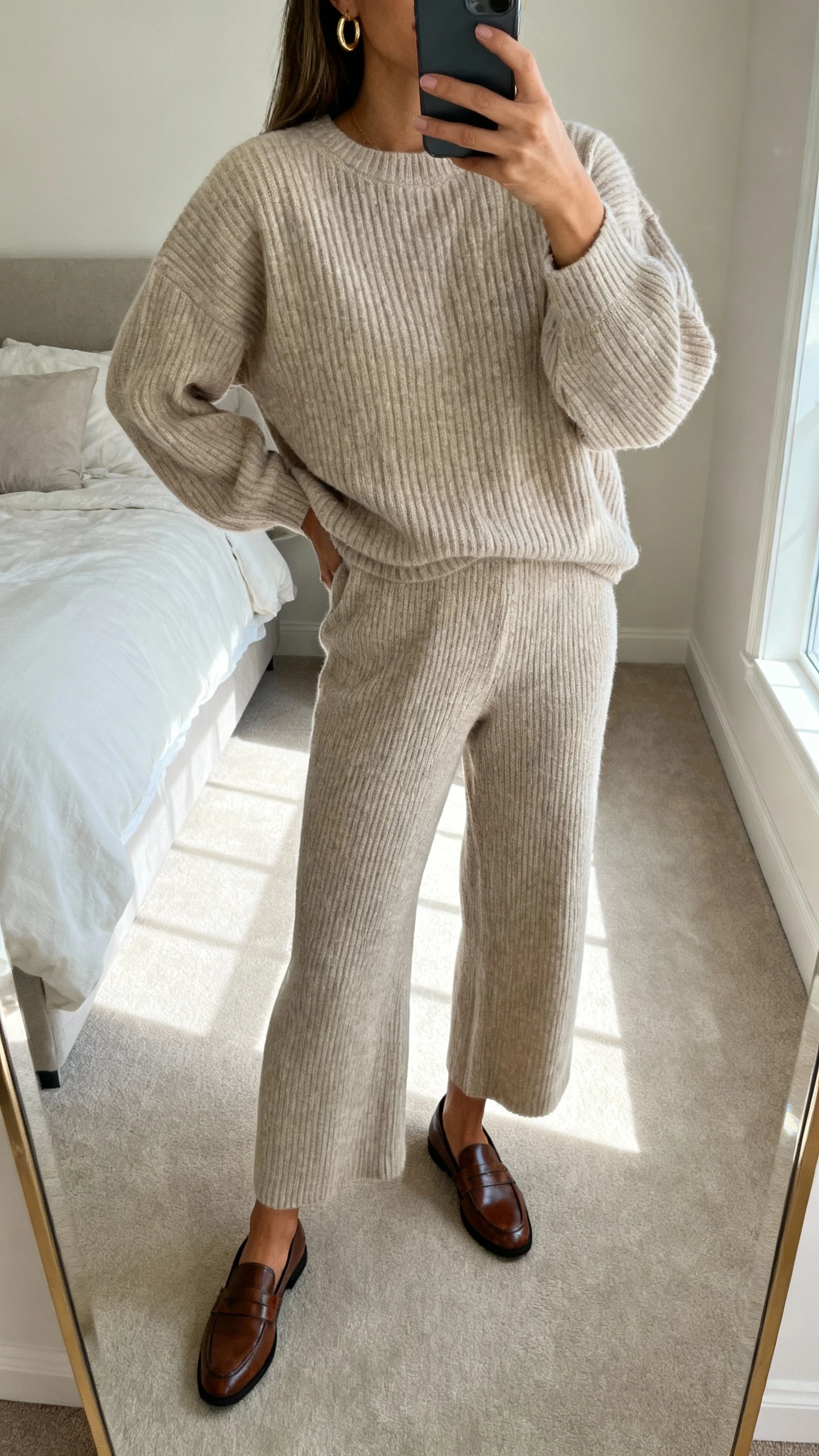 iPhone mirror selfie of a woman wearing an elevated knit set: matching ribbed knit sweater and knit wide-leg pants in a neutral tone, minimal gold hoops, sleek leather loafers, face not visible, cropped at neck, bedroom mirror with soft window light, natural iPhone photo quality.