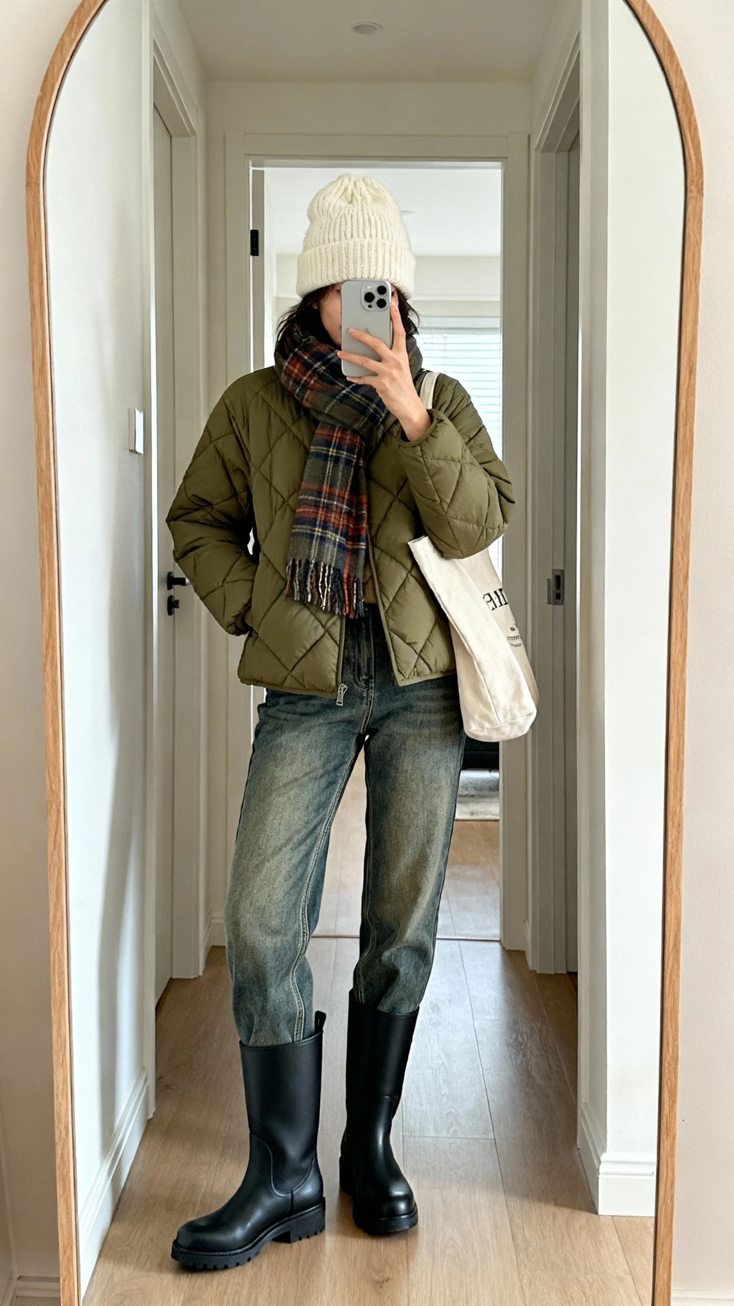 iPhone mirror selfie of a woman wearing an olive lightly padded quilted jacket, medium-wash mid-rise straight-leg denim, water-resistant black Chelsea boots, a cream chunky beanie, plaid cozy scarf, and a canvas tote; face not visible, hallway mirror, soft indoor daylight, natural iPhone photo quality.