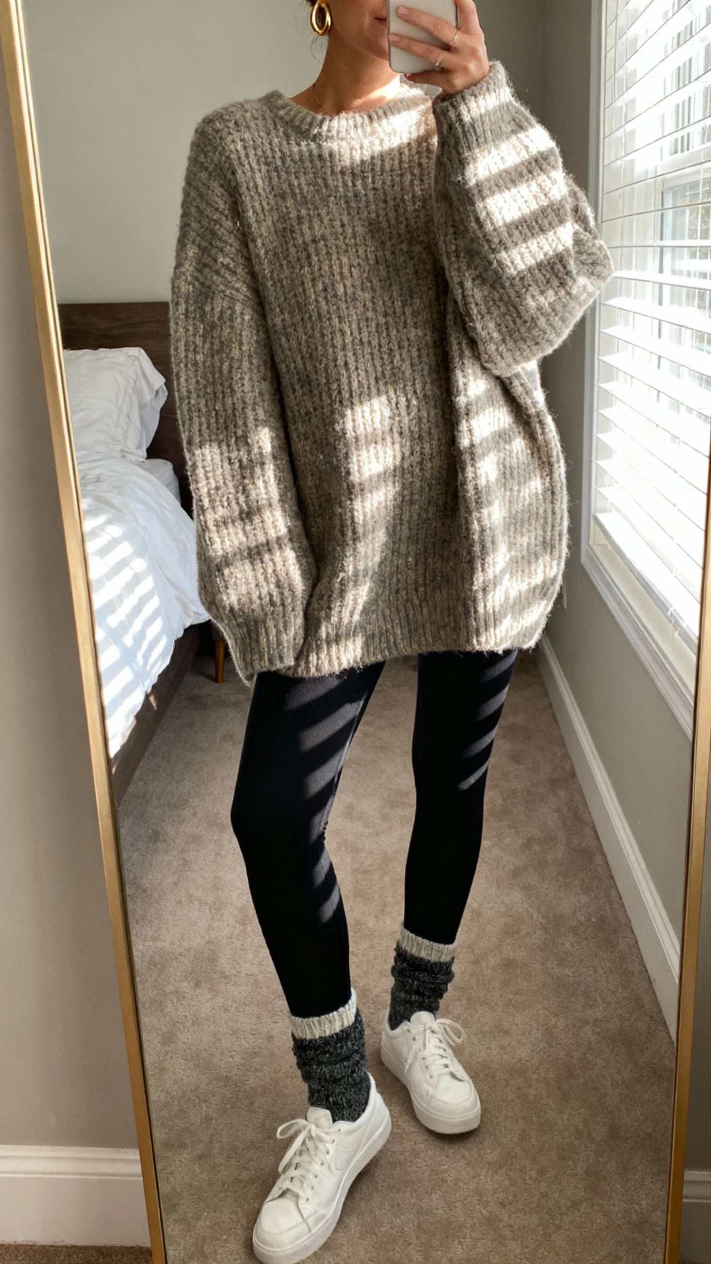 iPhone mirror selfie of a woman wearing an oversized chunky sweater, black leggings, cozy socks, white sneakers, simple gold hoops, face not visible, cropped at neck, bedroom with full-length mirror and morning window light, natural iPhone photo quality.