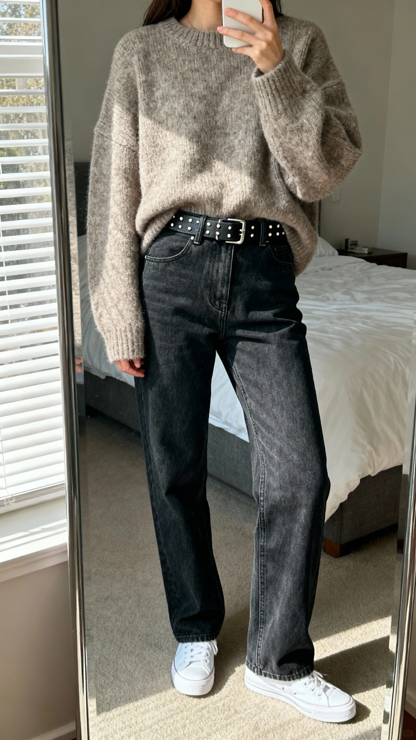 iPhone mirror selfie of a woman wearing an oversized sweater with straight-leg jeans, classic belt, simple studs, white sneakers, face not visible, cropped at neck, bedroom mirror with natural window light, natural iPhone photo quality.
