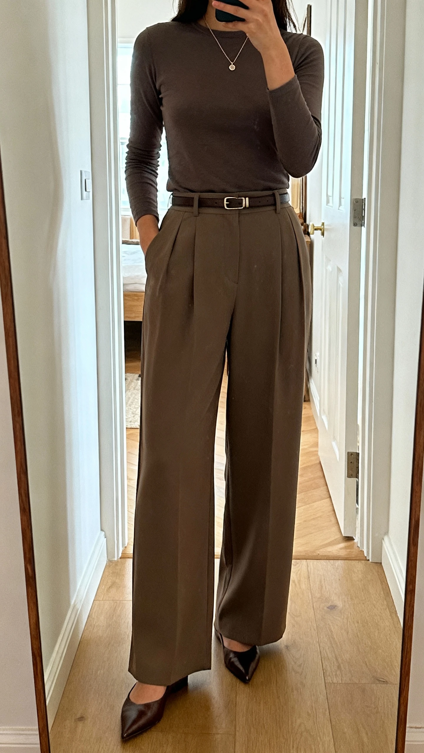 iPhone mirror selfie of a woman wearing high-waisted wide-leg trousers with a fitted top, pointed flats, slim belt, delicate pendant, face not visible, full-length mirror in entryway, natural daylight, natural iPhone photo quality