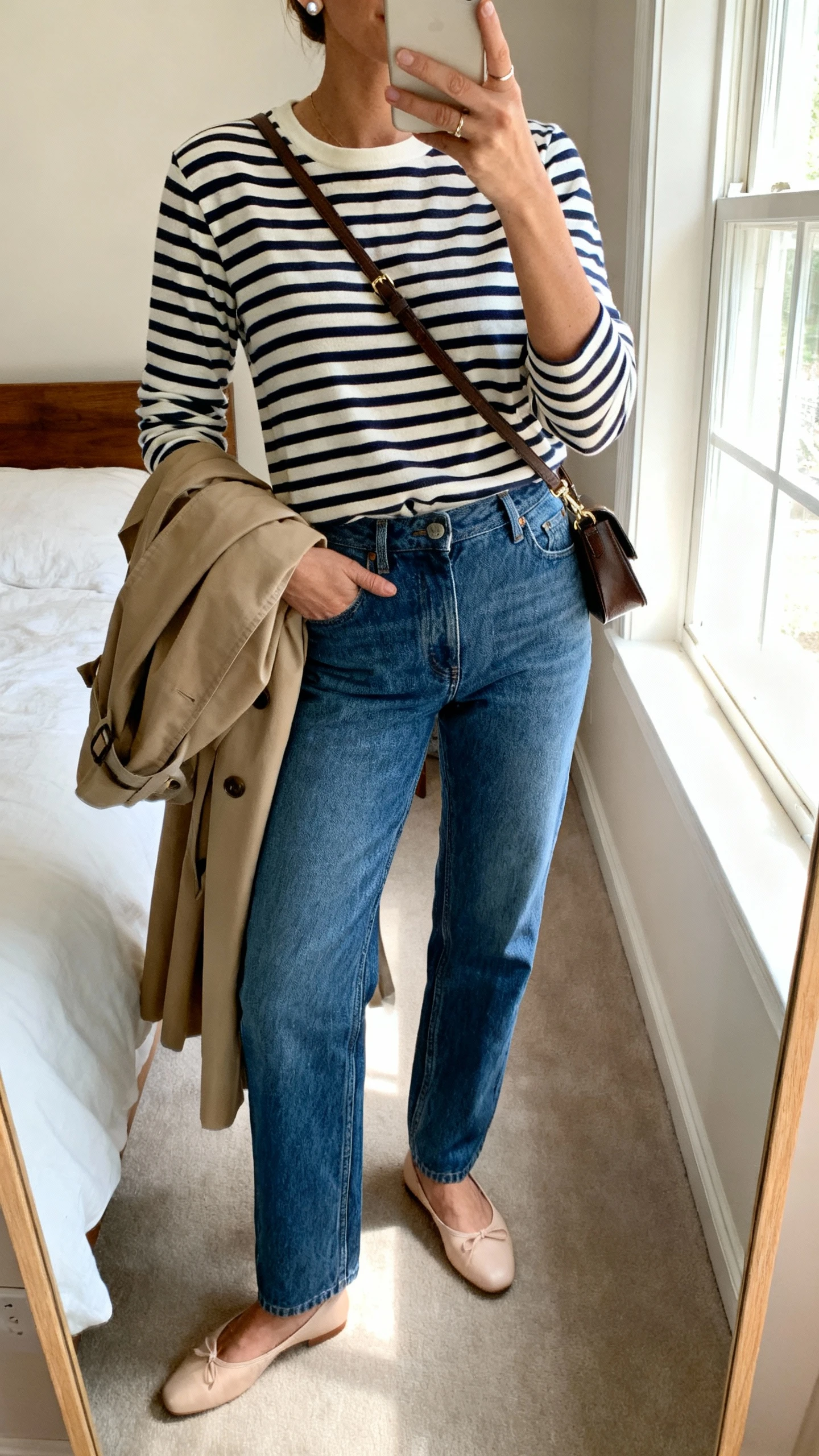 iPhone mirror selfie of a woman wearing Parisian-off-duty stripes: classic navy-and-cream striped long-sleeve tee, high-rise straight blue jeans, ballet flats, trench tossed over arm, crossbody bag, simple studs, face not visible, cropped at shoulders, bedroom mirror with window light, casual iPhone photo