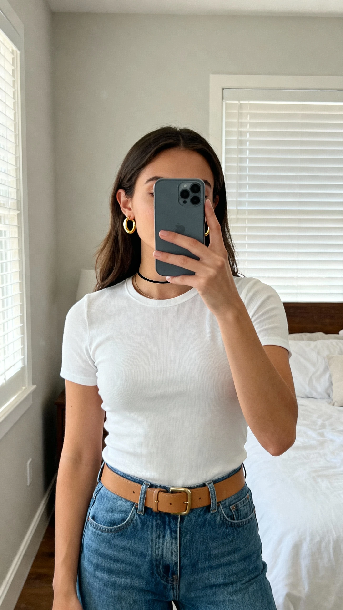 iPhone mirror selfie of a woman wearing simple clean gold hoop earrings, white fitted tee, high-waisted blue jeans, tan belt, minimal makeup, holding phone by neck so face not visible, bedroom mirror with natural window light, natural iPhone photo quality