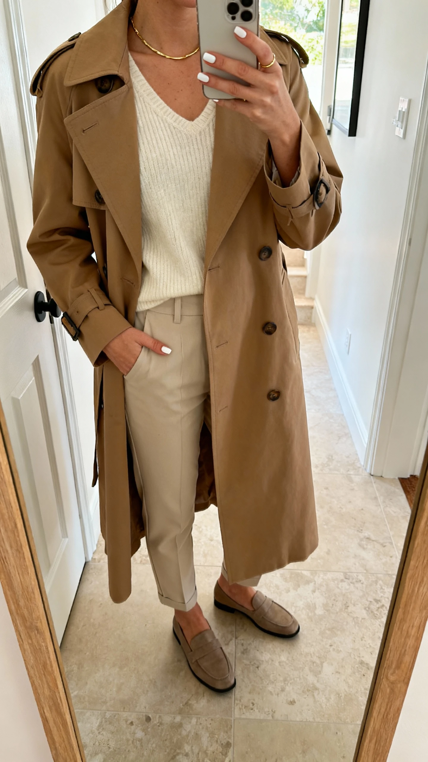 iPhone mirror selfie of a woman with beige base nails and soft white micro half-moons, wearing a camel trench, cream knit, tapered trousers, loafers, thin gold necklace, face not visible, entryway mirror, natural daylight, casual iPhone photo.