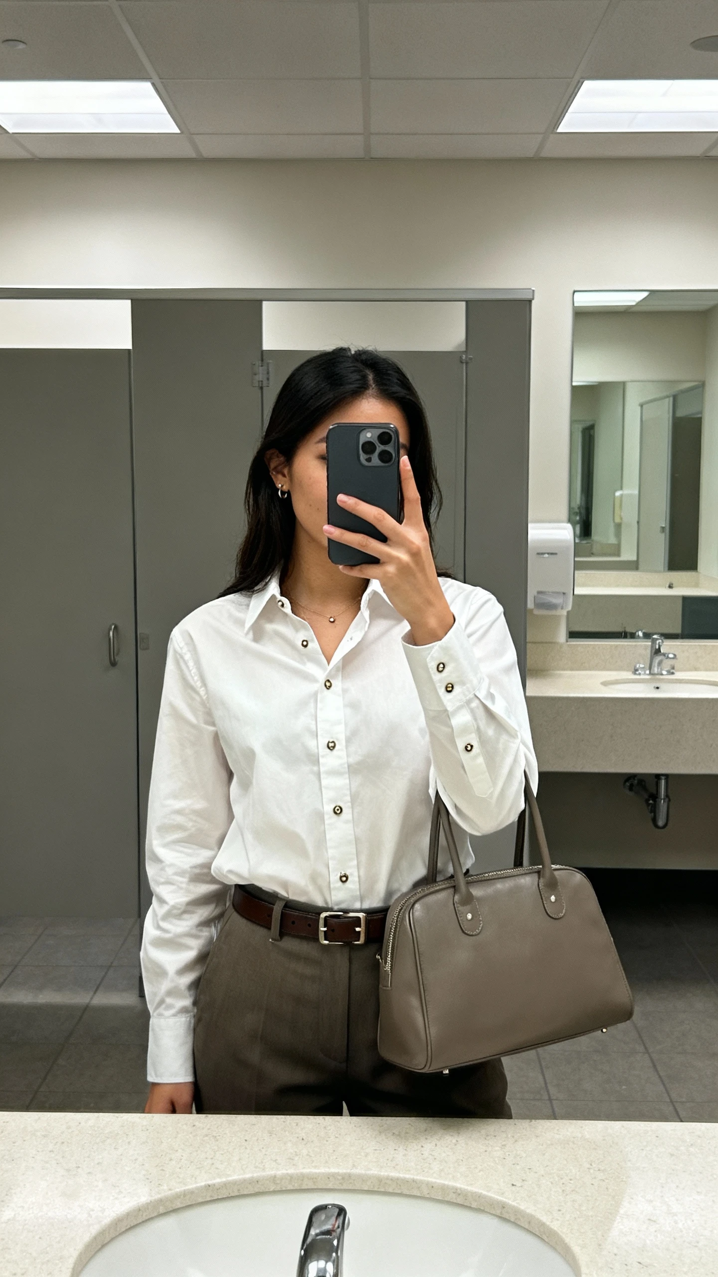 iPhone office-bathroom mirror selfie of a woman wearing a crisp button-up shirt tucked into trousers, slim leather belt, simple studs, structured tote, face not visible, indoor lighting, natural iPhone photo quality