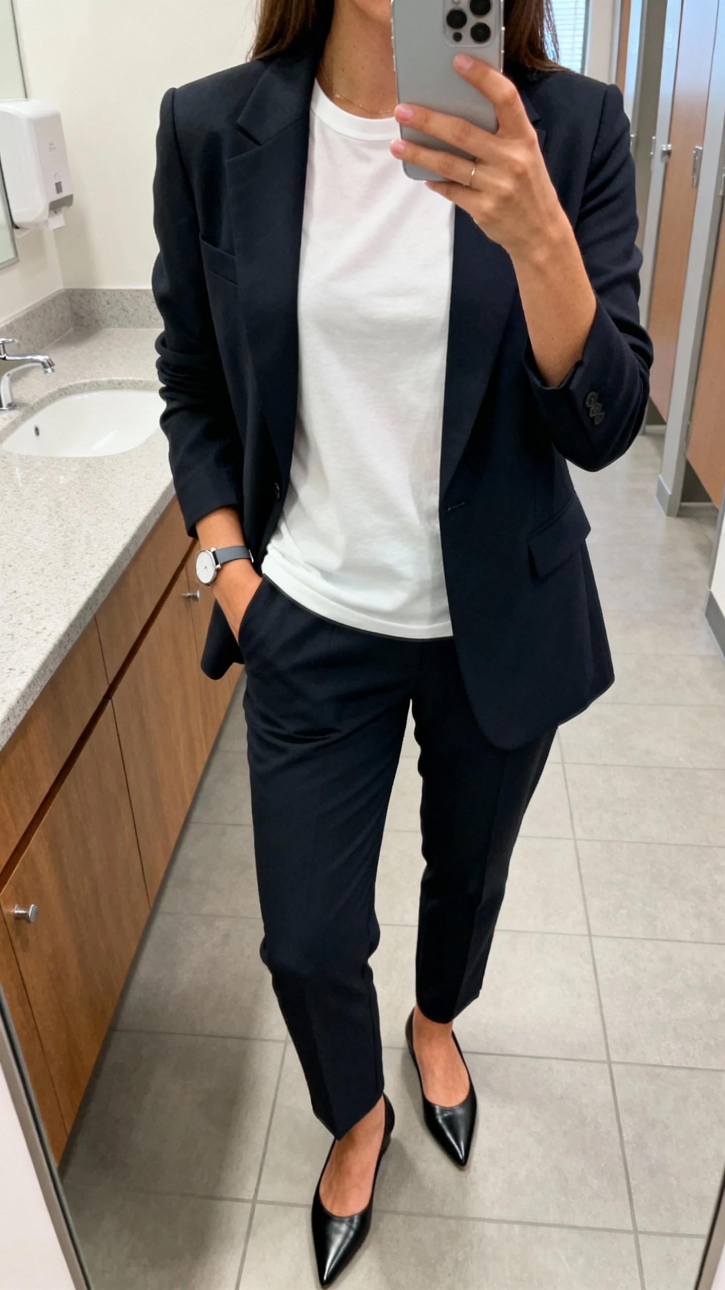 iPhone office mirror selfie of a woman in a tailored blazer over a white crew-neck T-shirt, tailored trousers, pointed flats, minimalist watch, face not visible, cropped at shoulders, bright office bathroom lighting, natural iPhone aesthetic.