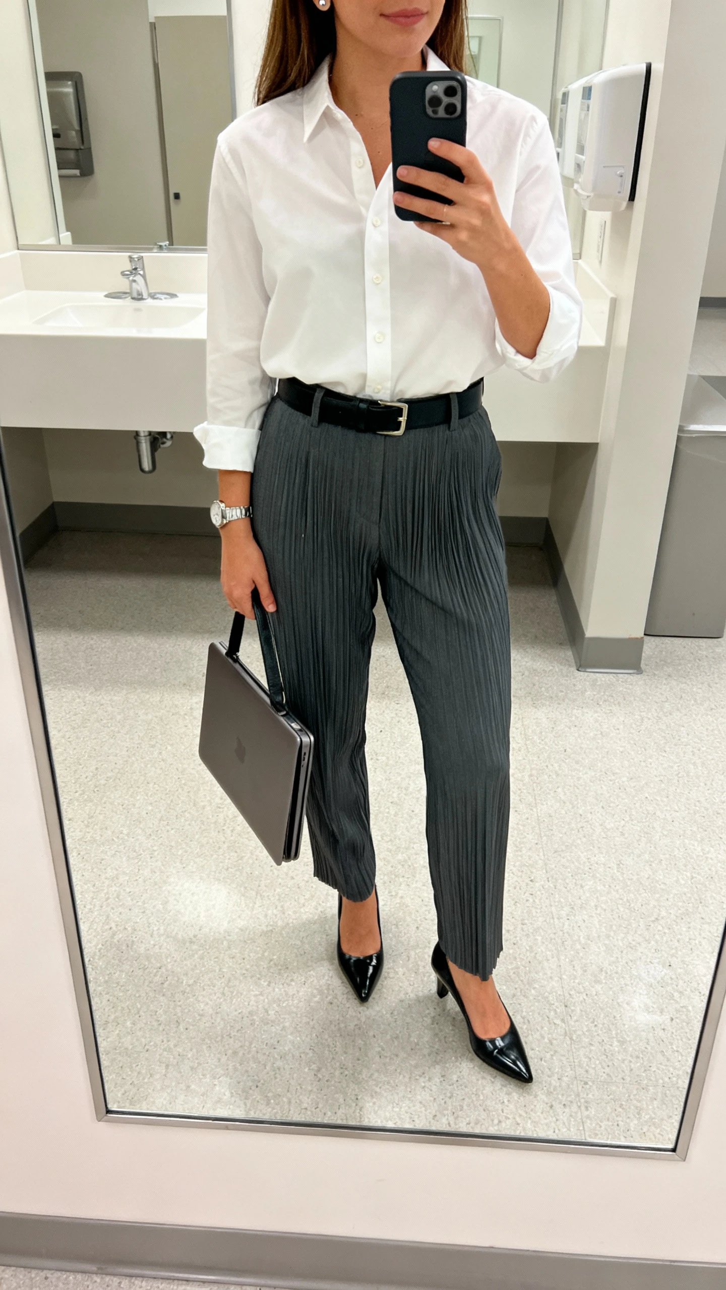 iPhone office mirror selfie of a woman wearing a crisp white button-down tucked into charcoal pleated trousers, black belt, polished black pumps, slim leather laptop bag, simple stud earrings and a watch; office restroom mirror, face not visible, natural indoor lighting, iPhone photo quality.