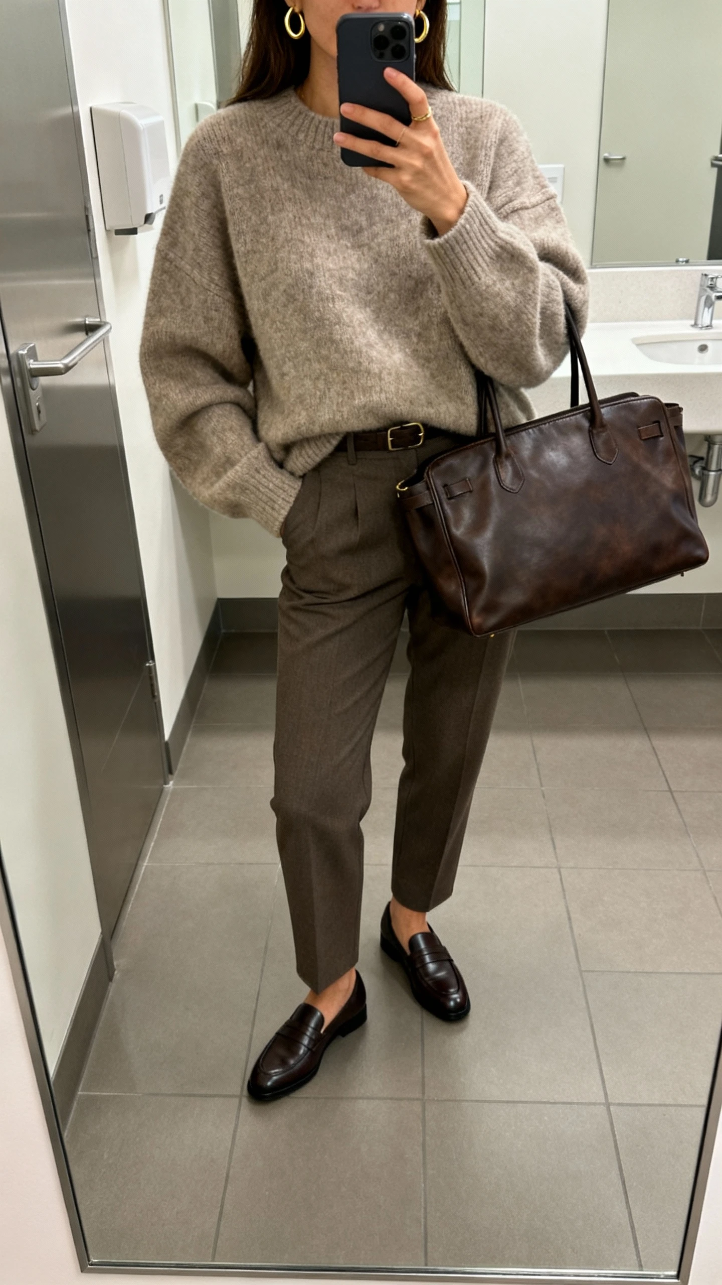 iPhone office restroom mirror selfie of a woman in an oversized cashmere sweater tucked into tailored trousers, sleek loafers, minimal gold hoops, and a structured leather tote, face not visible, cropped at shoulders, soft indoor lighting, natural iPhone photo quality.