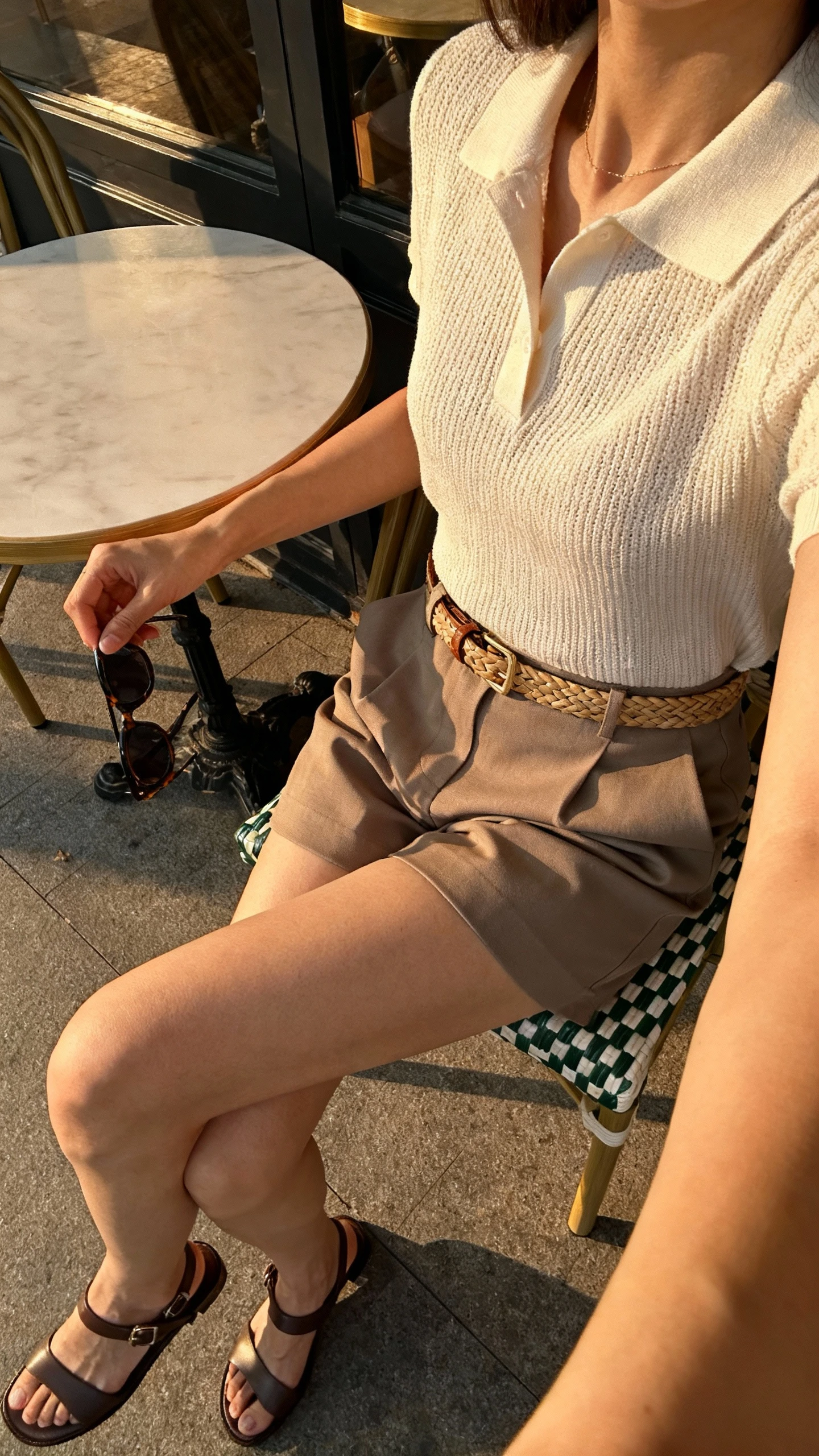 iPhone patio selfie side angle of a woman wearing a knit polo tucked into tailored shorts, sleek sandals, woven belt, sunglasses in hand, face not visible, late afternoon natural light at a cafe table, natural iPhone photo quality.