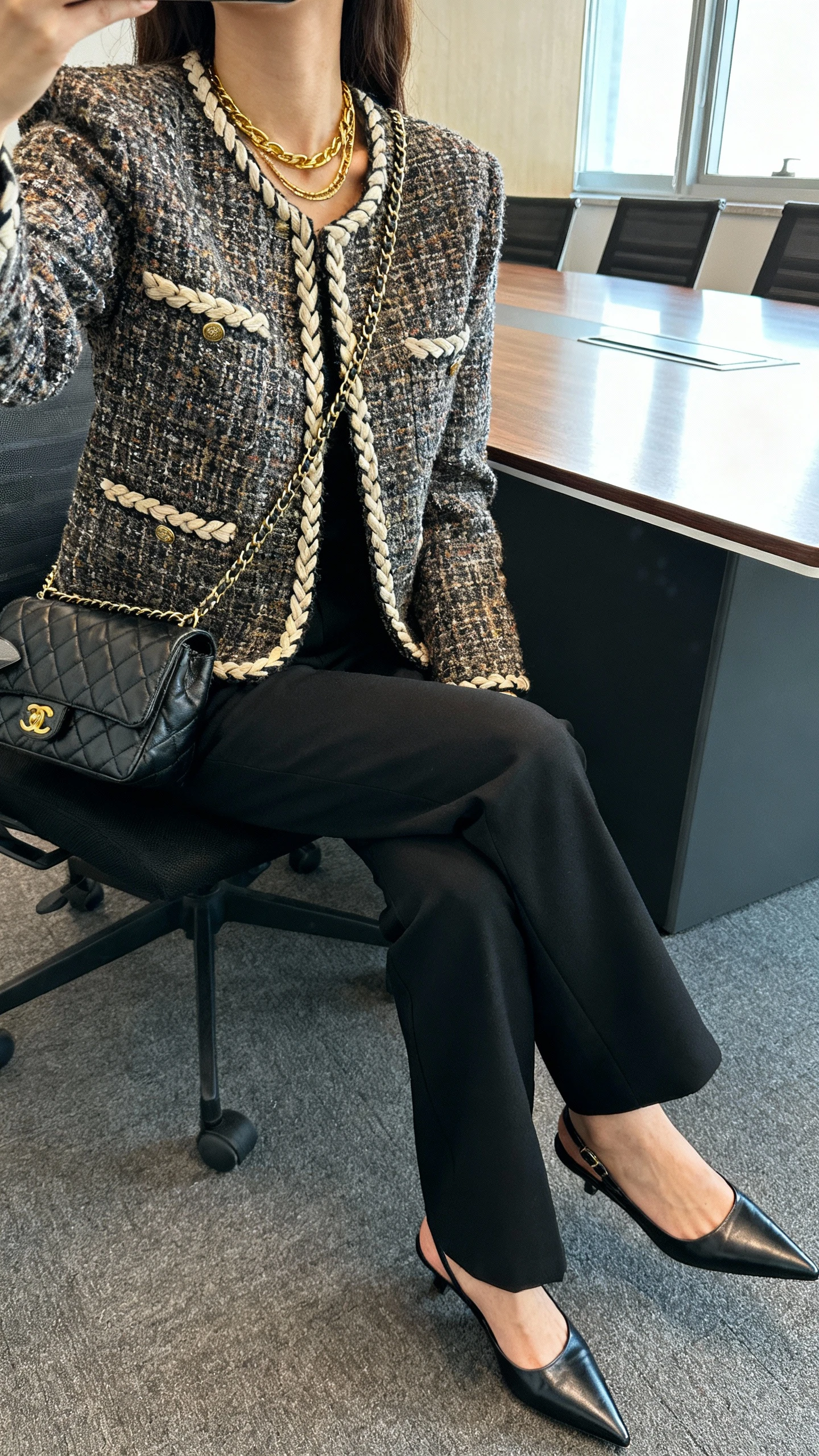iPhone seated selfie cropped at shoulders showing a woman wearing a tweed jacket with braided trim, tailored black trousers, pointed slingbacks, quilted chain-strap bag, layered gold necklaces; face not visible, office conference room table, daylight from window, natural iPhone photo quality.