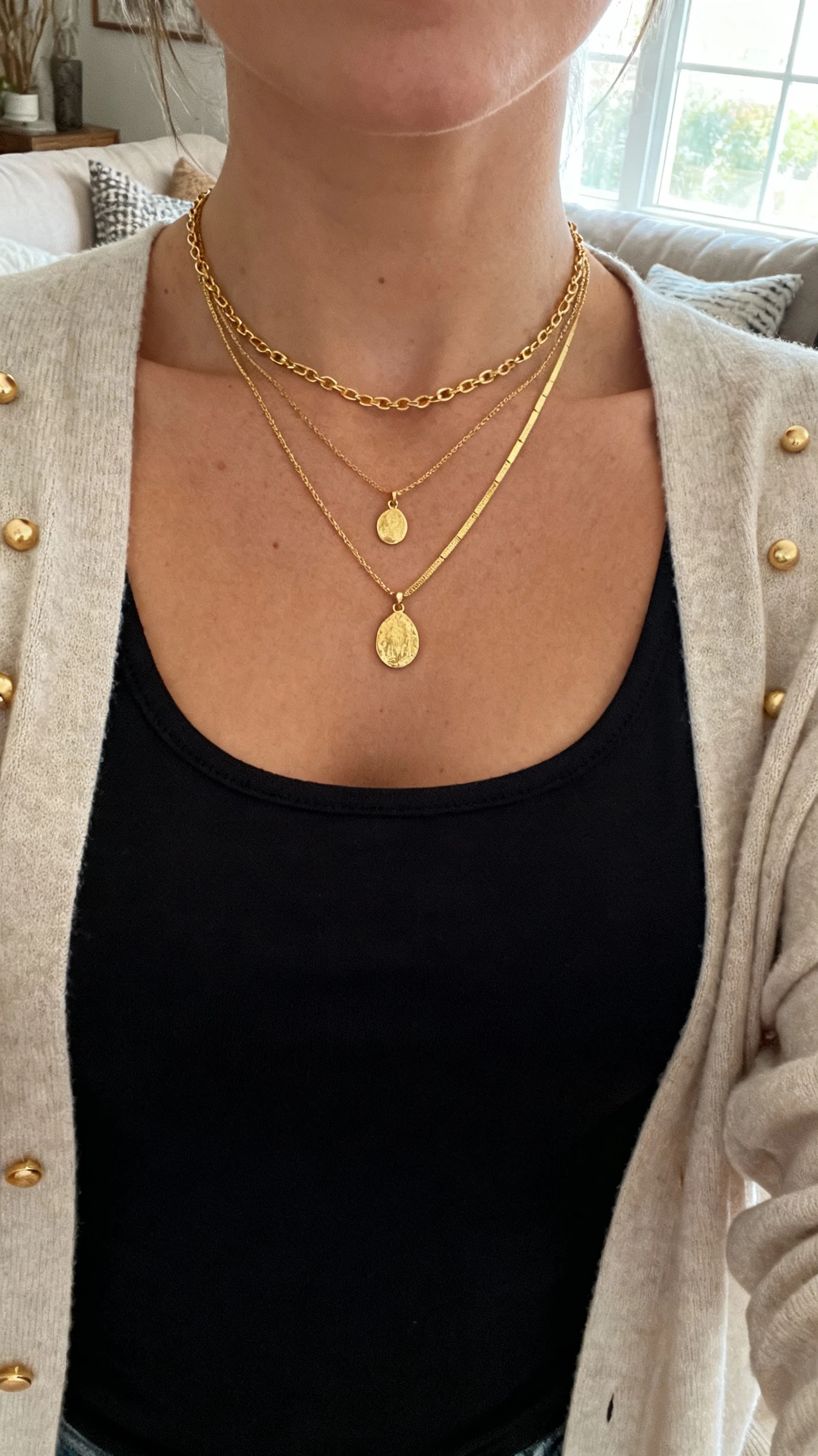 iPhone selfie cropped at collarbone showing a woman layering delicate gold necklaces (short chain + mid-length pendant + longer fine chain) over a black scoop-neck top, with small gold studs and light cardigan, face not visible, cozy living room near window, natural iPhone photo quality