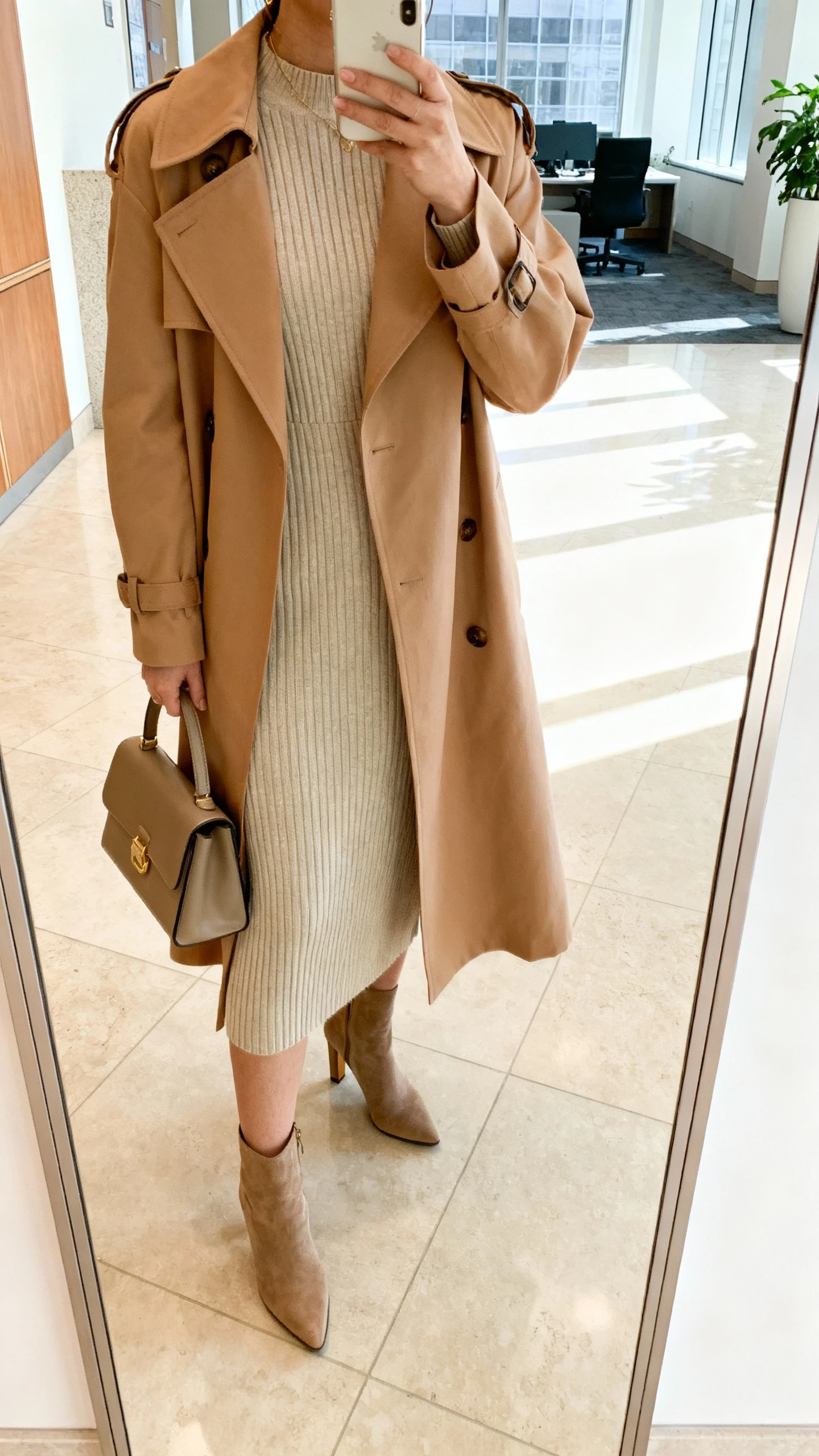 iPhone selfie cropped at neck showing a woman in a soft camel trench layered over a ribbed knit dress, heeled ankle boots, and a structured handbag, face not visible, office lobby mirror with bright indoor lighting, natural iPhone photo.