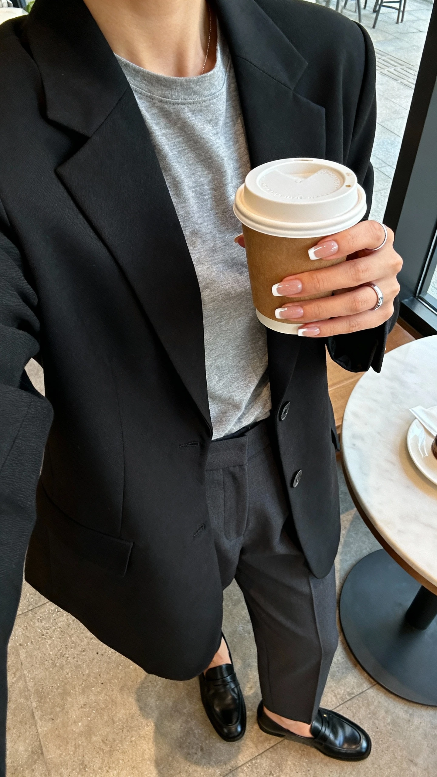 iPhone selfie cropped at shoulders of a woman holding a coffee cup to show modern skinny French line nails, wearing a black blazer over a gray tee, tailored trousers, delicate silver ring, and black loafers; cafe table setting, morning light, face not visible, natural iPhone aesthetic.