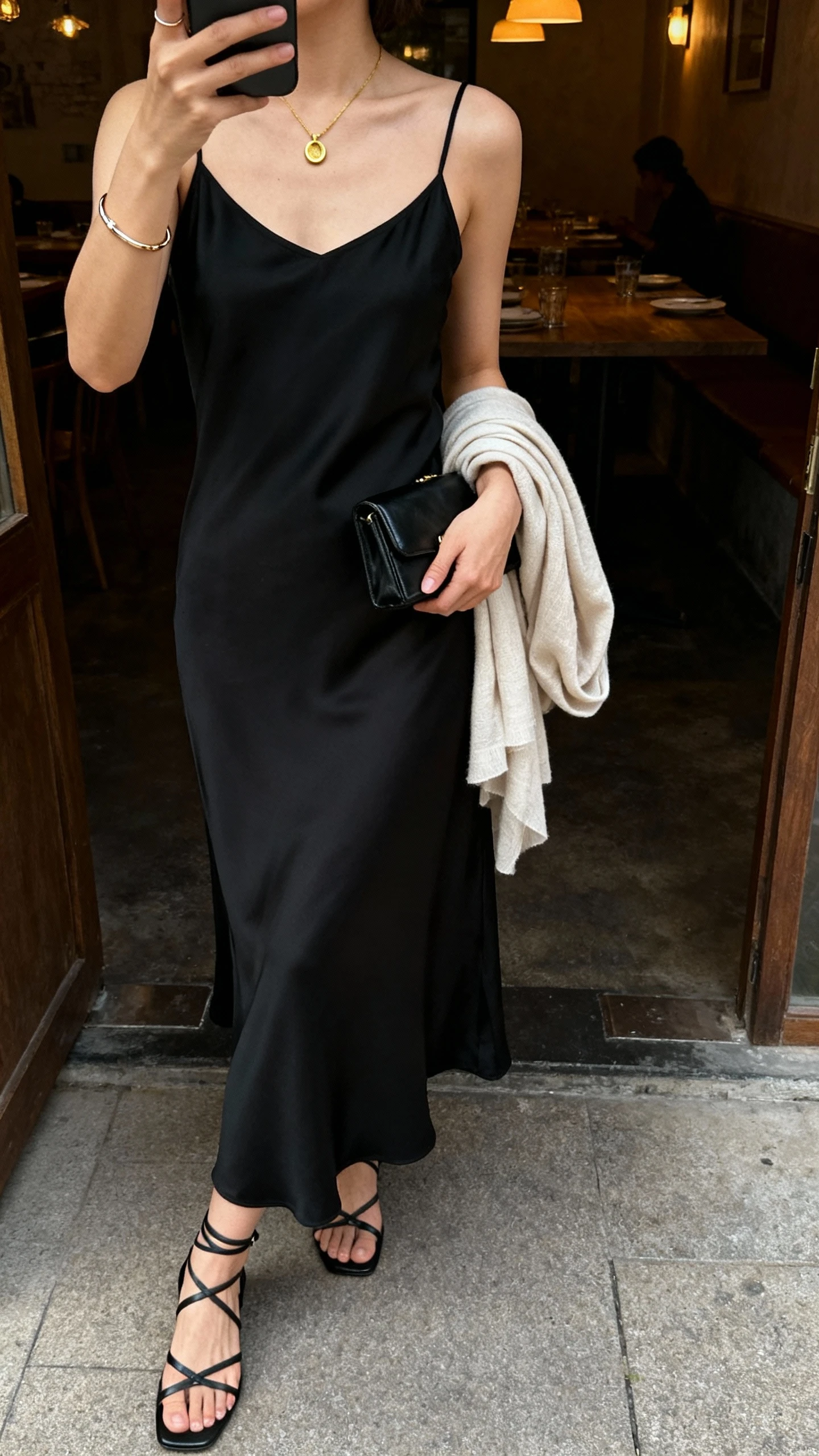 iPhone selfie cropped at shoulders showing a woman in a sleek black slip dress, minimal strappy heels, fine gold pendant, slim bracelet, small black clutch, light shawl draped over arm, face not visible, dim restaurant entryway with warm indoor lighting, natural iPhone photo quality.