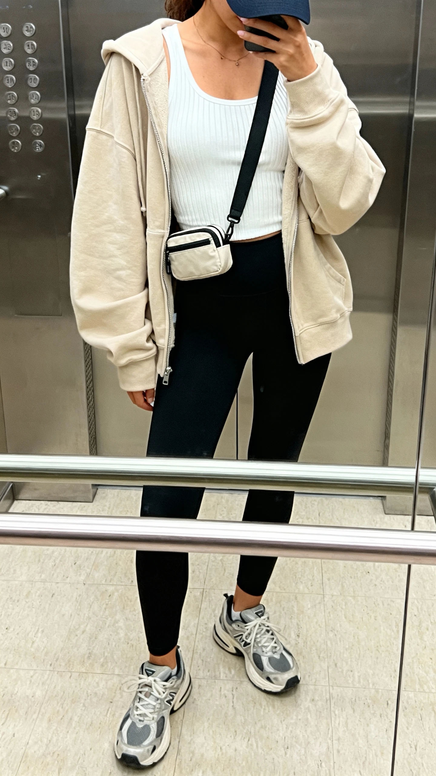 iPhone selfie cropped at shoulders showing a woman in a sporty-chic look: black leggings, white ribbed tank, oversized beige zip-up hoodie, chunky dad sneakers, baseball cap, small belt bag worn crossbody, face not visible, elevator mirror, afternoon indoor lighting, natural iPhone photo quality.