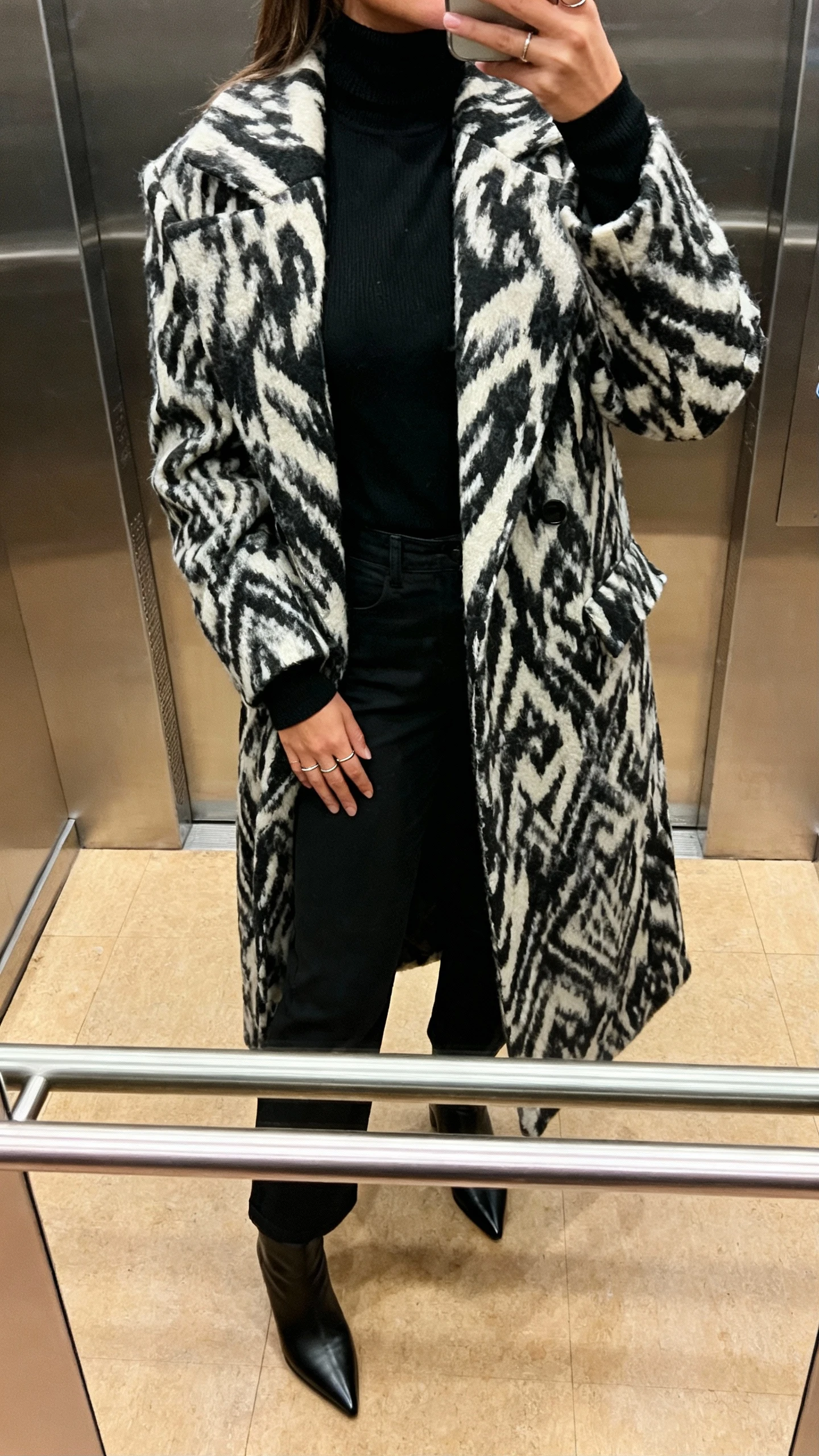 iPhone selfie cropped at shoulders showing a woman in a column of black (black turtleneck and black trousers) with a bold statement coat, pointed boots and minimal rings, face not visible, elevator mirror, warm indoor lighting, natural iPhone photo quality.
