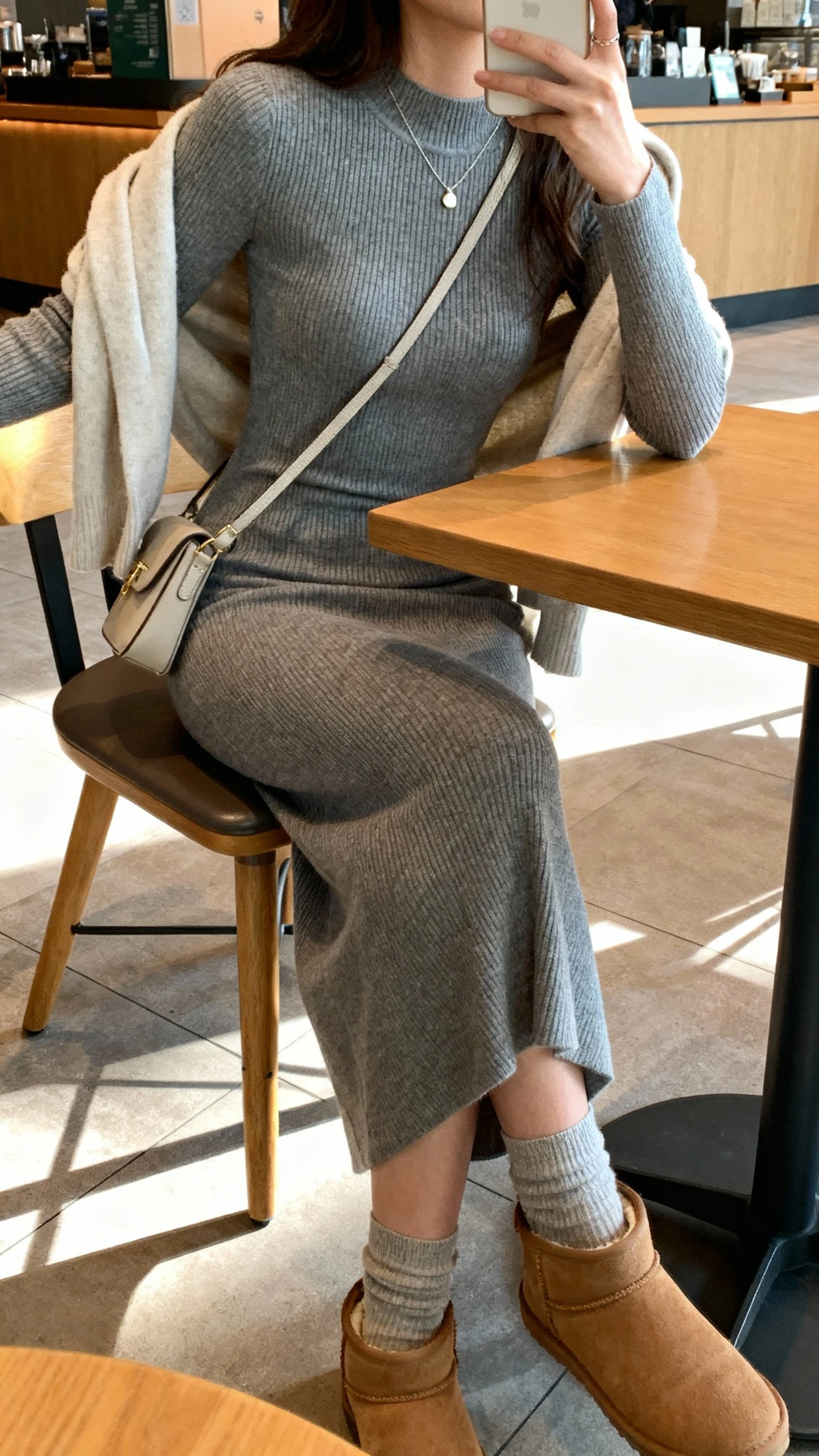 iPhone selfie side angle of a woman in a soft knit dress: body-skimming ribbed knit midi dress in heather gray, minimal pendant necklace, ankle sock boots, small crossbody, light cardigan draped over shoulders, face not visible, seated at a coffee shop table with warm indoor light, natural iPhone photo quality.