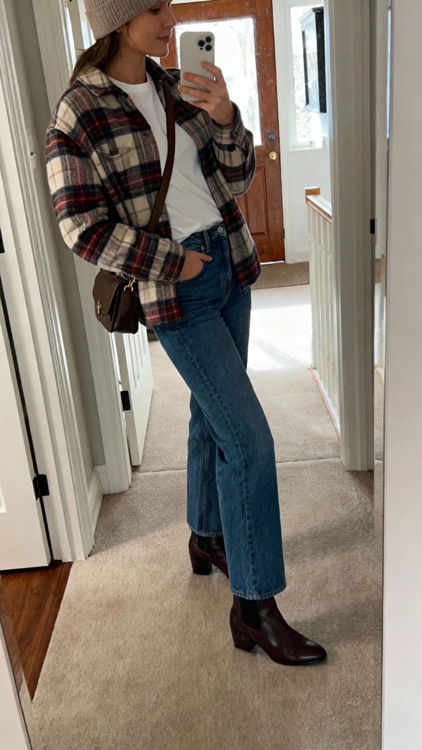 iPhone side-angle hallway mirror selfie of a woman wearing a plaid shacket over a white tee, straight-leg blue jeans, ankle boots, crossbody bag, beanie, face not visible, natural daylight by entryway, casual iPhone photo.