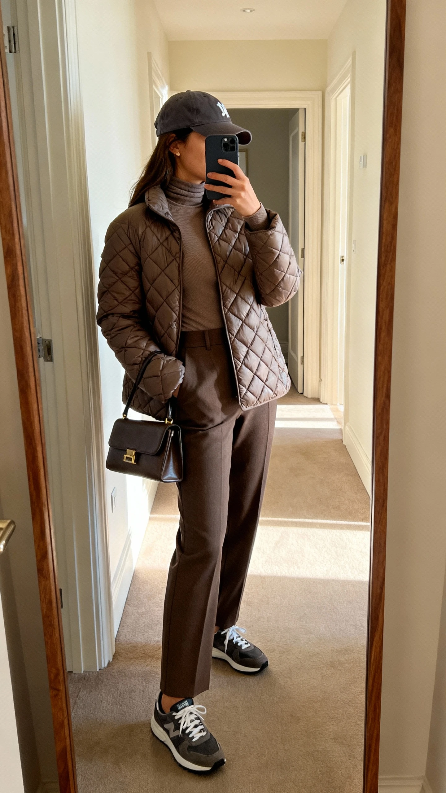 iPhone side-angle hallway mirror selfie of a woman in a quilted puffer jacket layered over a fitted turtleneck, tailored trousers, sleek sneakers, small structured tote, and a baseball cap, face not visible, indoor afternoon light, casual iPhone photo.