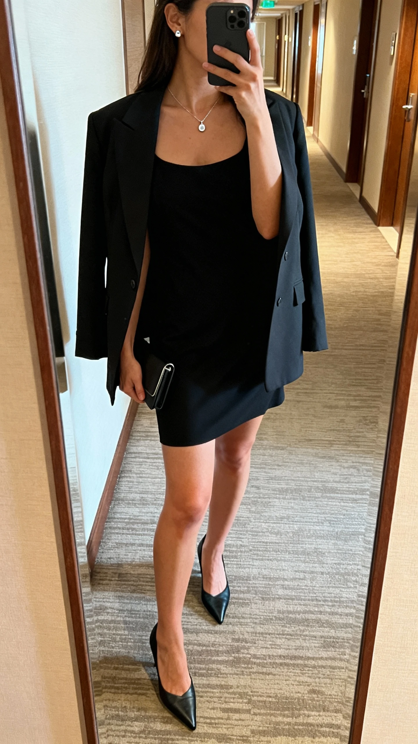 iPhone side-angle mirror selfie of a woman in a little black dress with pointed heels, structured blazer over shoulders, delicate pendant necklace, stud earrings, minimal clutch in hand; face not visible, hotel hallway mirror, warm indoor lighting, casual iPhone aesthetic.