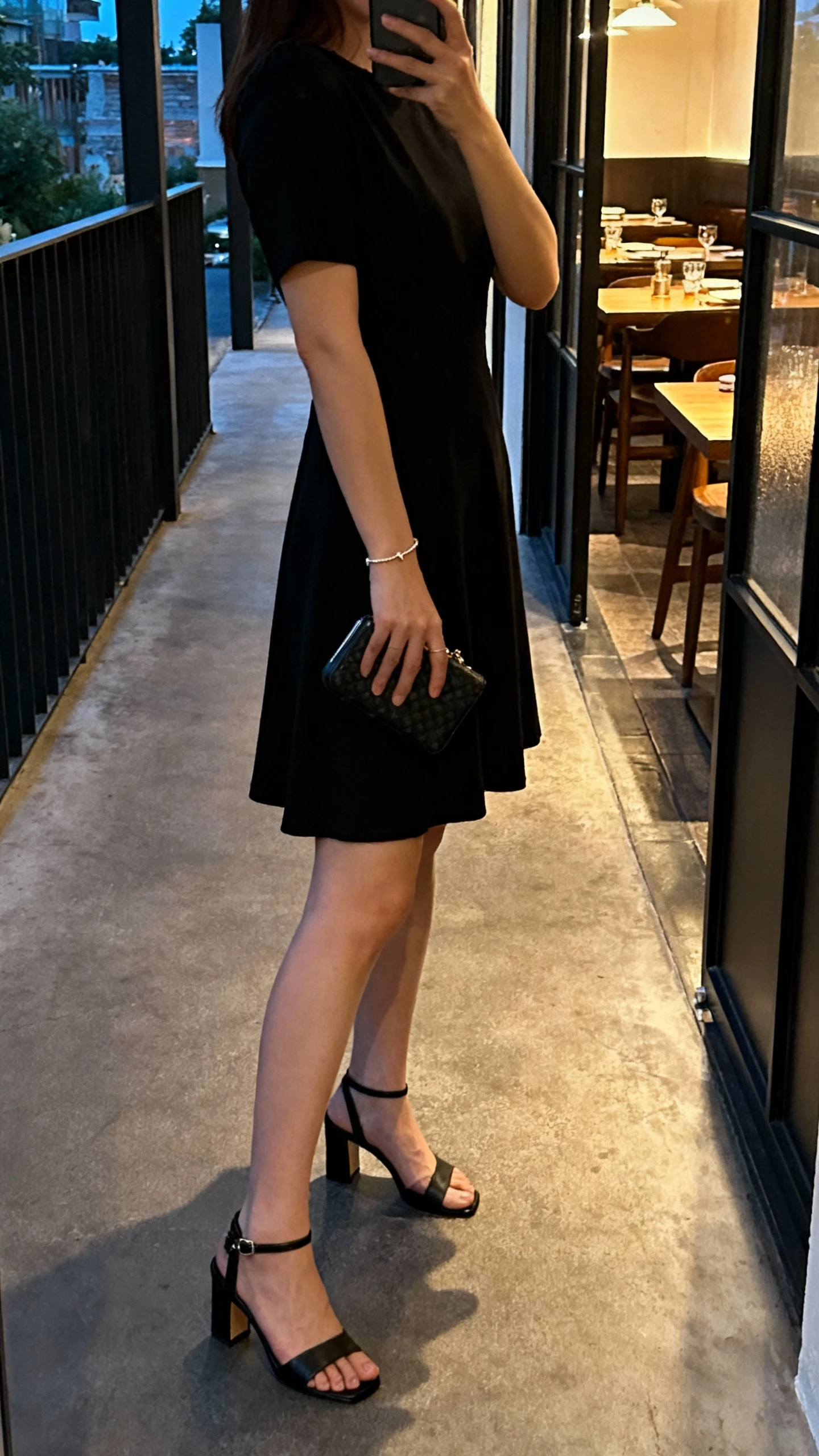 iPhone side-angle selfie of a woman in a classic little black dress with black heeled sandals, small clutch, delicate bracelet, face not visible, hallway outside a restaurant, warm evening indoor lighting, natural iPhone aesthetic.