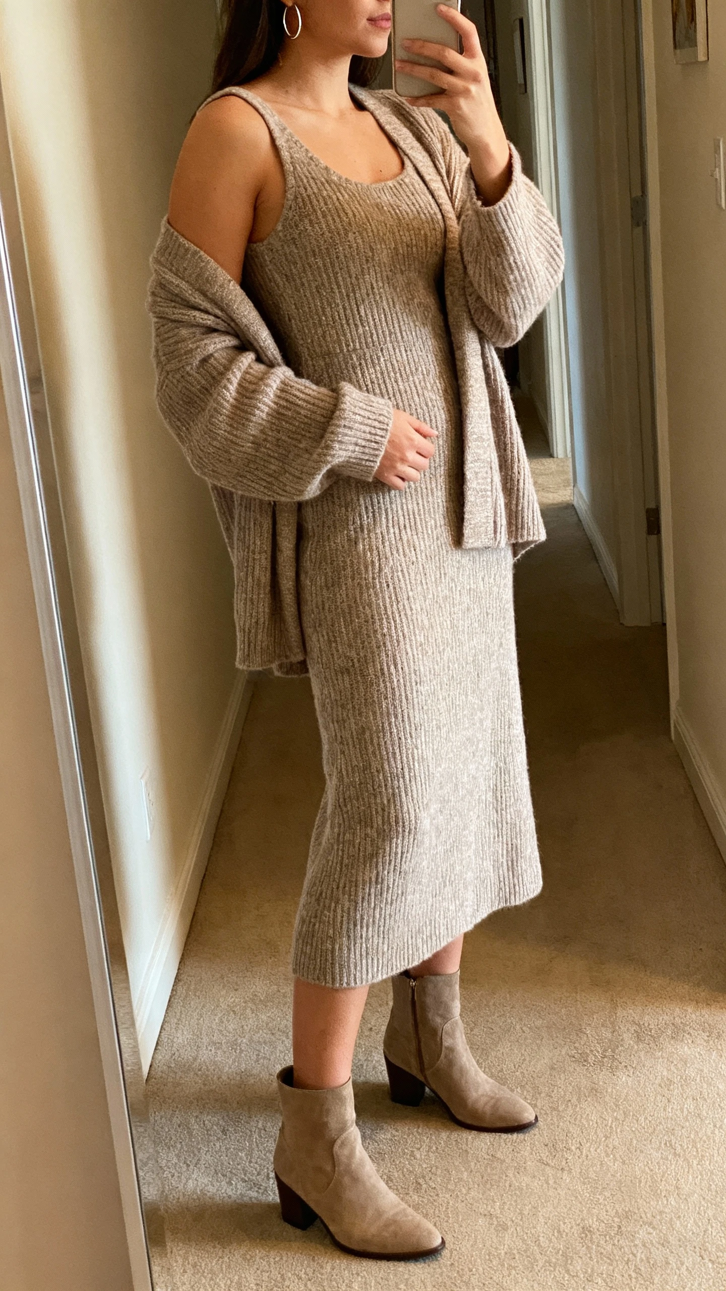 iPhone side-angle selfie of a woman wearing a matching knit dress set (sleeveless midi knit dress with a coordinating cardigan), ankle boots and minimal hoops, face not visible, hallway mirror with warm indoor lighting, natural iPhone photo quality.