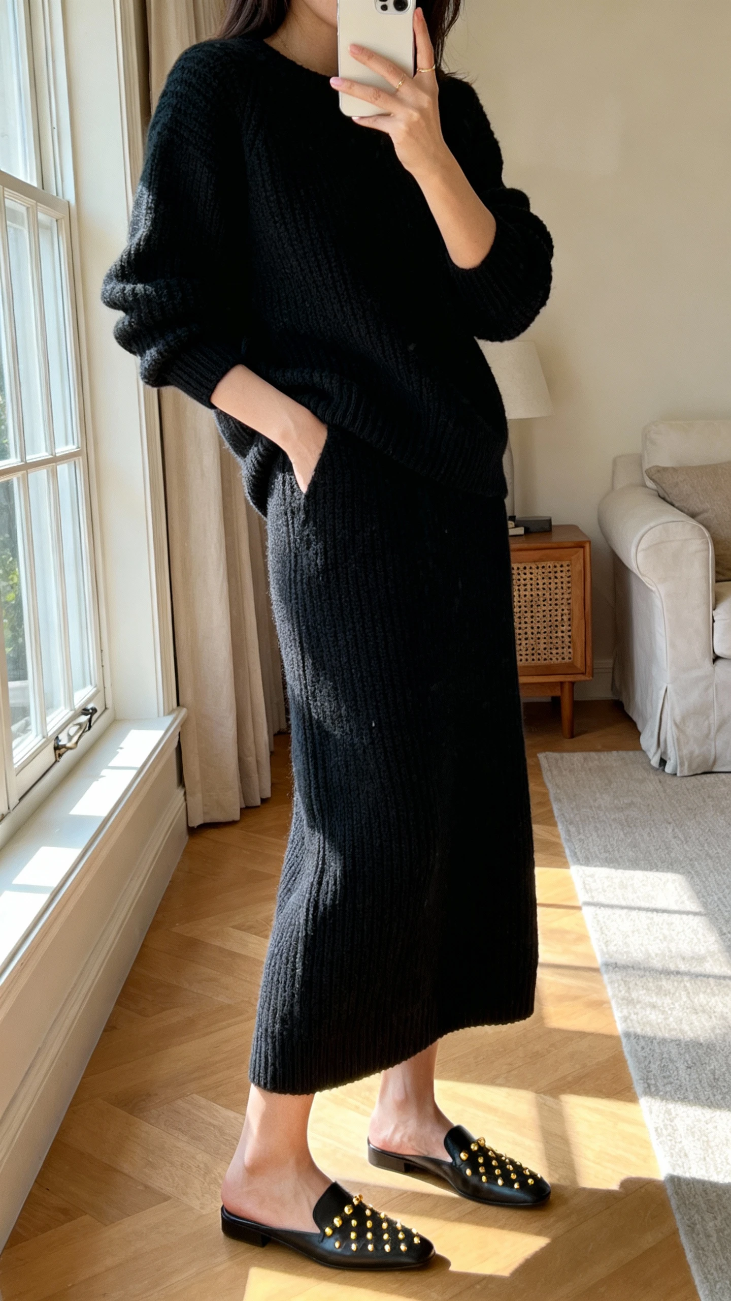 iPhone side-angle selfie of a woman wearing a monochrome knit set (matching sweater and knit skirt) with sleek flats and subtle gold studs, face not visible, living room near window, morning light, natural iPhone photo quality.