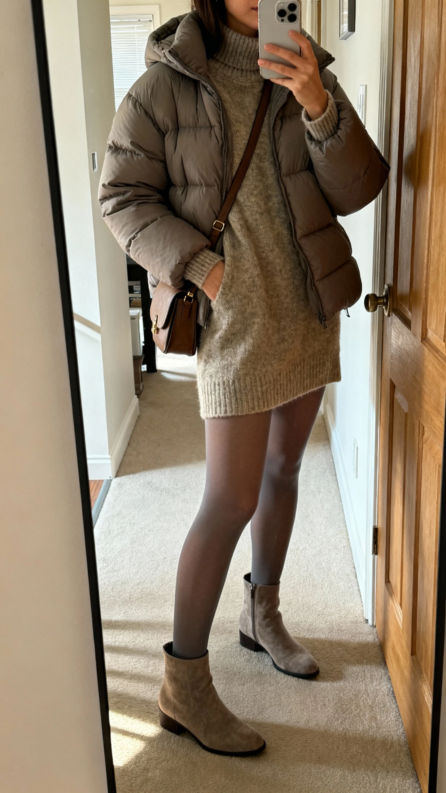 iPhone side-angle selfie of a woman wearing an elevated puffer jacket over a sweater dress, with opaque tights, ankle boots, and a crossbody bag, face not visible, hallway mirror near apartment door, morning light, natural iPhone photo quality.