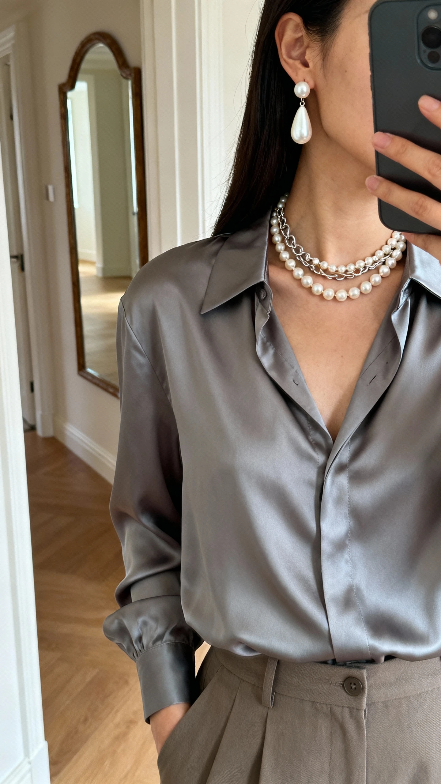iPhone side-angle selfie showing a woman wearing modern pearl jewelry (asymmetric pearl drop earrings and a pearl-chain mix necklace) with a sleek satin blouse and tailored trousers, face not visible, hallway mirror, soft indoor lighting, natural iPhone photo quality
