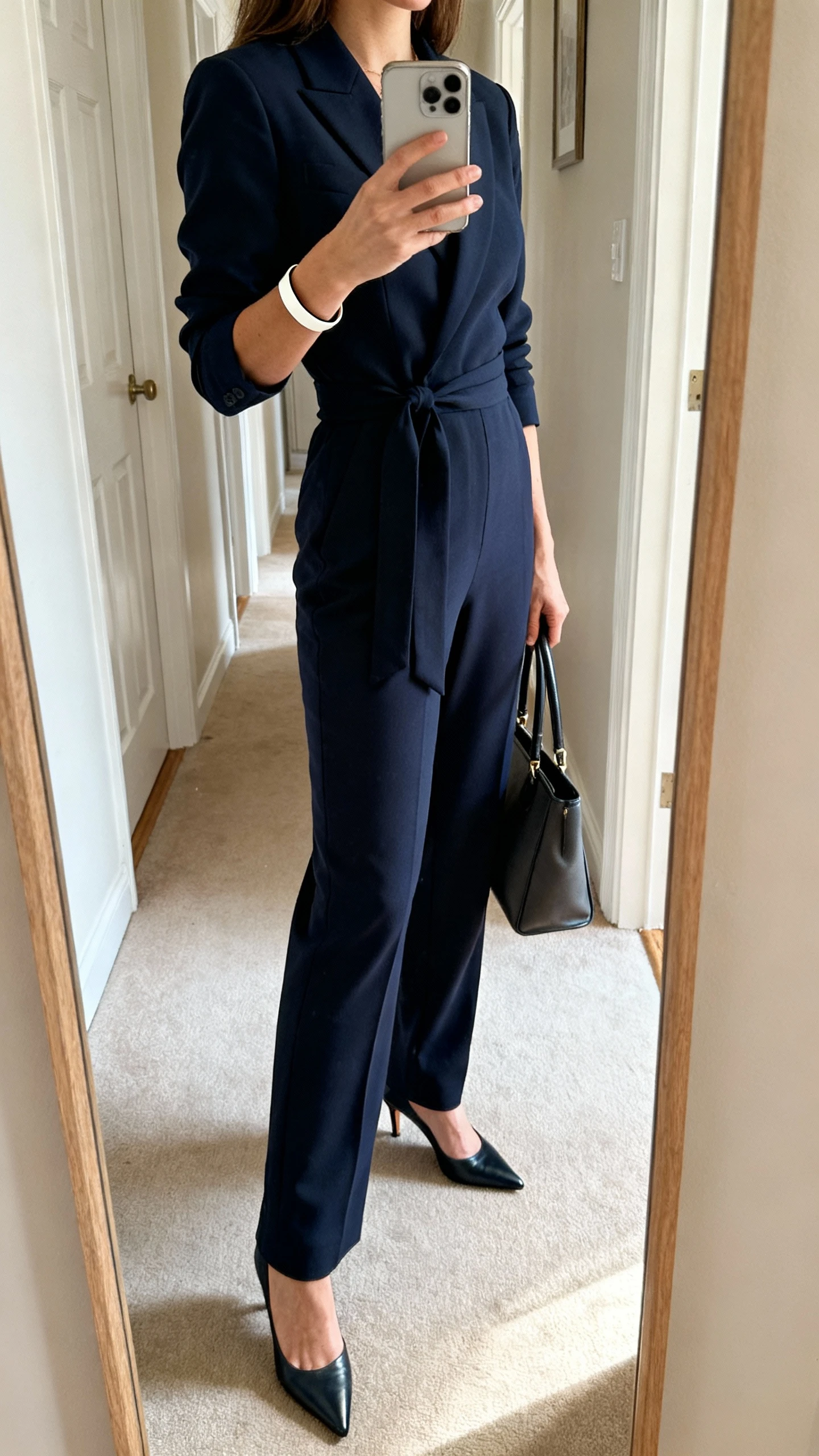 iPhone side-profile mirror selfie of a woman in a sharp tailored jumpsuit (navy), cinched at the waist, with pointed heels, minimalist cuff bracelet, and a top-handle bag; hallway mirror, face not visible, indoor daylight, natural iPhone look.