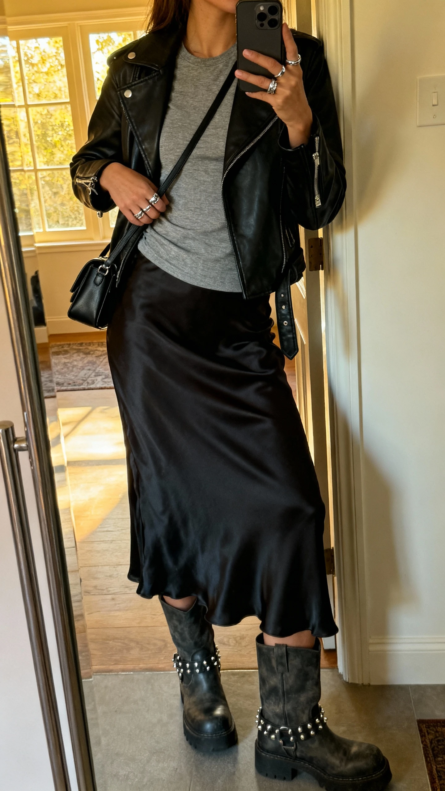 iPhone side-view selfie of a woman in a minimal slip skirt + leather jacket: black leather moto jacket over a silky black slip skirt, fitted gray tee, chunky boots, silver studs and rings, small black shoulder bag, face not visible, entryway mirror at home with afternoon window light, natural iPhone photo quality.