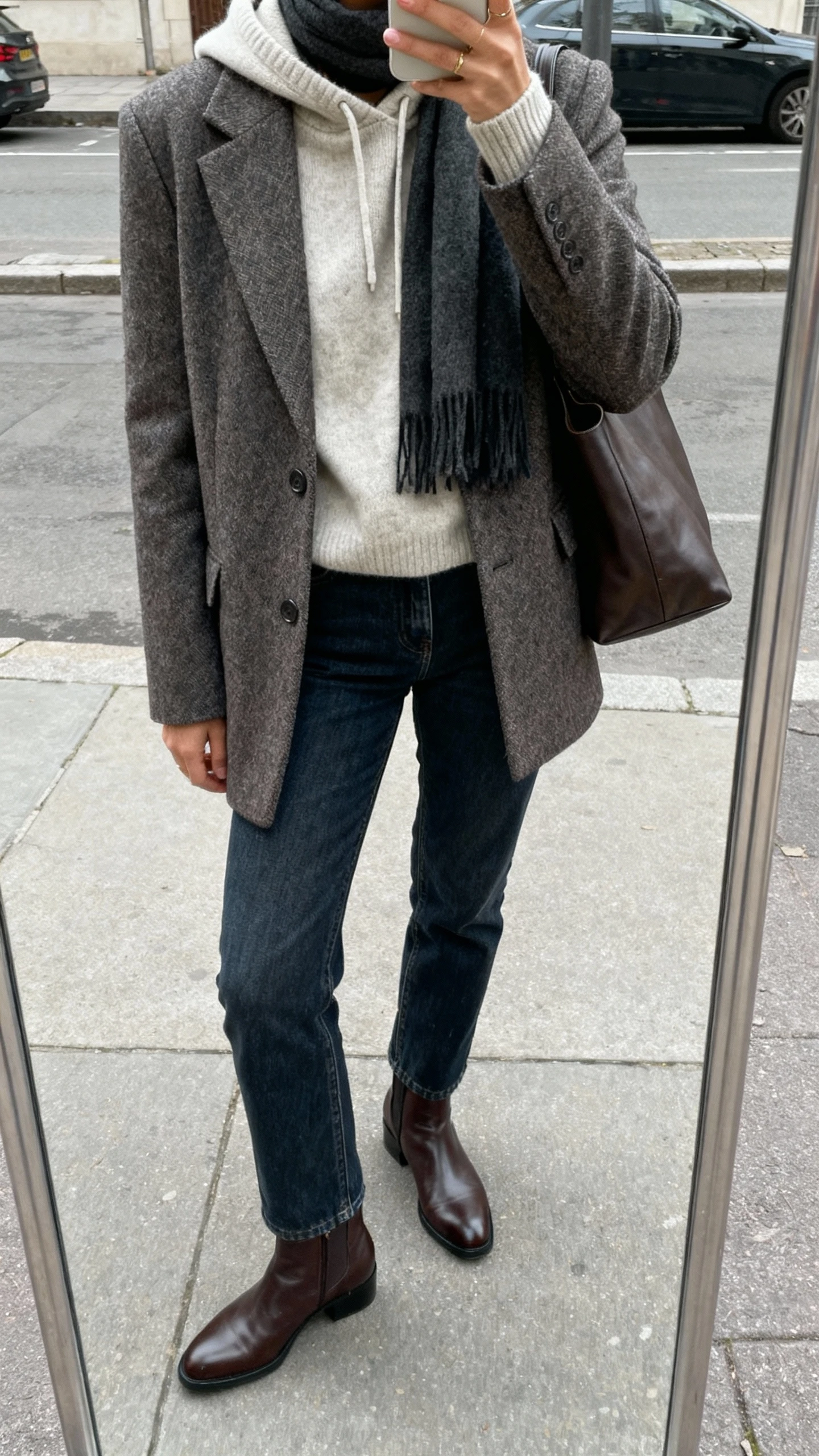 iPhone street-mirror selfie of a woman wearing a cashmere hoodie under a tailored wool coat, straight-leg jeans, leather ankle boots, scarf and tote, face not visible, cropped at neck, overcast daylight, natural iPhone photo quality.