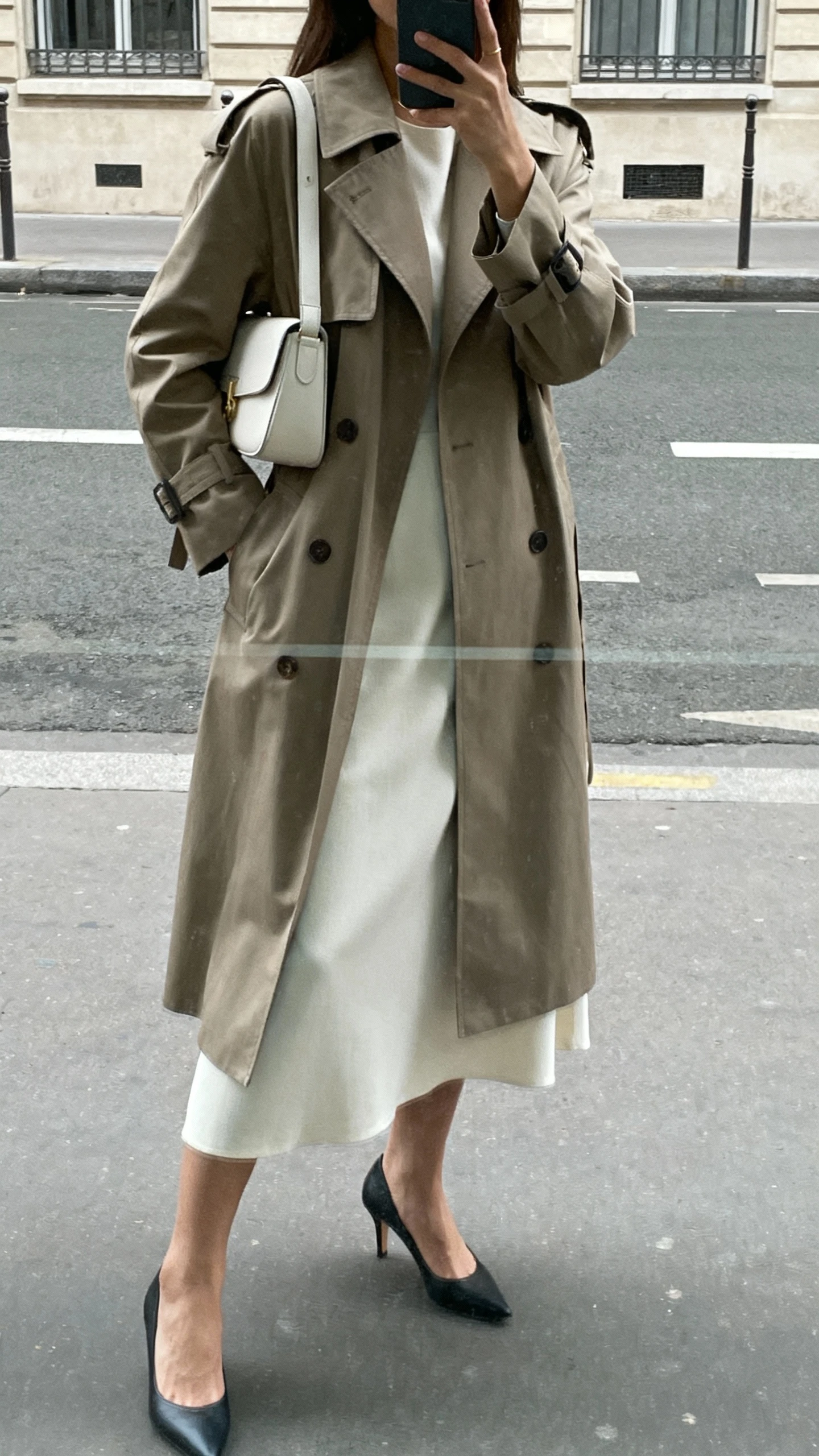 iPhone street reflection selfie of a woman wearing a minimalist dress layered under a classic trench coat, sleek low heels, clean shoulder bag, face not visible, overcast daylight, natural iPhone photo quality
