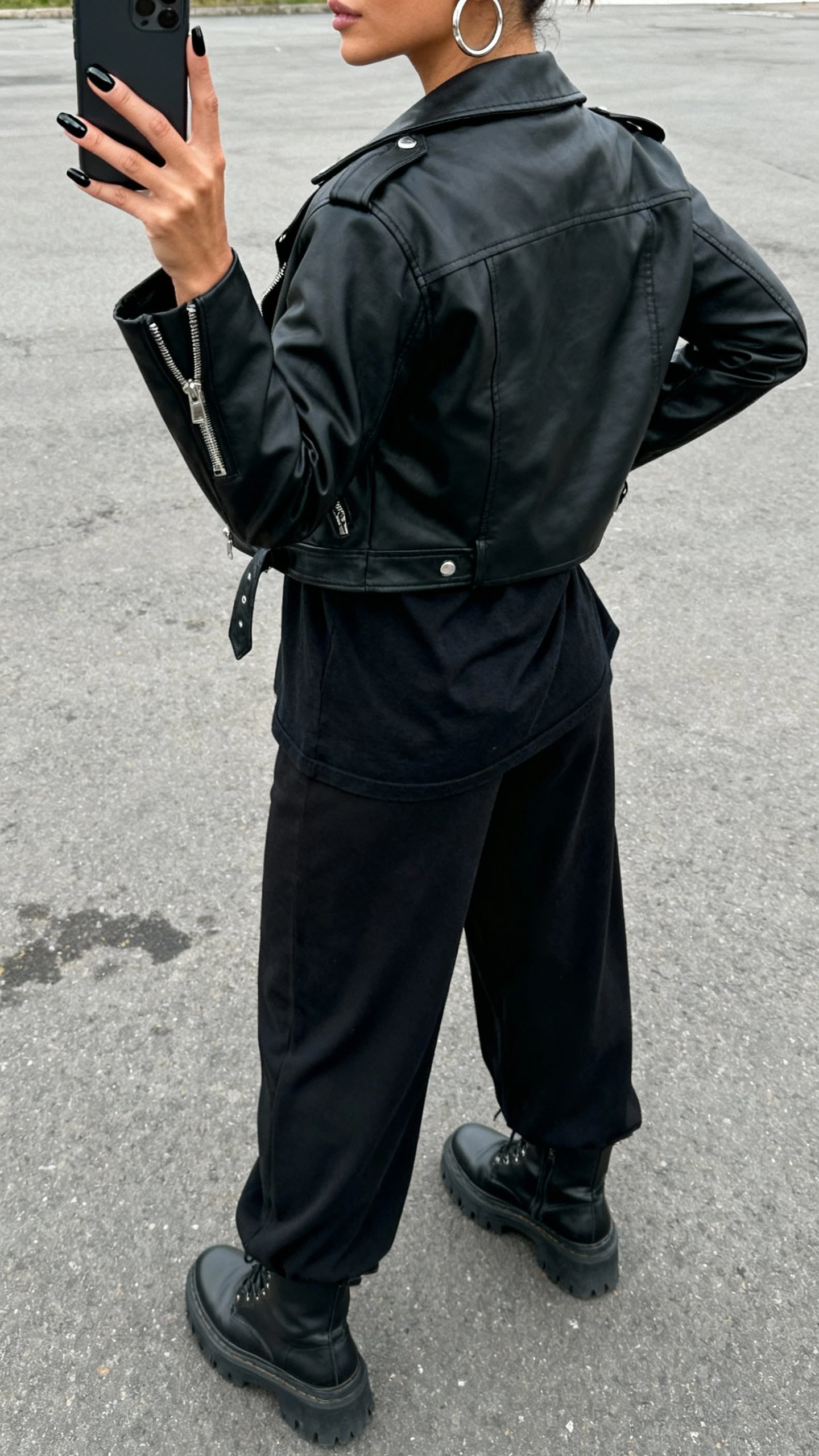 iPhone street-side back-view selfie of a woman with glossy black nails, wearing a black moto jacket, black tee, black wide-leg pants, chunky boots, silver hoops, face not visible, over-the-shoulder angle, overcast daylight, natural iPhone aesthetic.