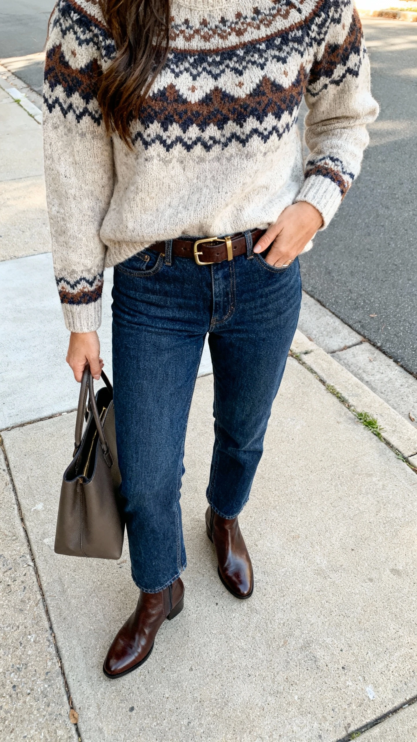 iPhone street-style selfie from chest down showing a woman in a Fair Isle sweater, straight-leg denim, polished leather ankle boots, a belt, and a structured tote, face not visible, sidewalk in soft afternoon daylight, natural iPhone photo quality.