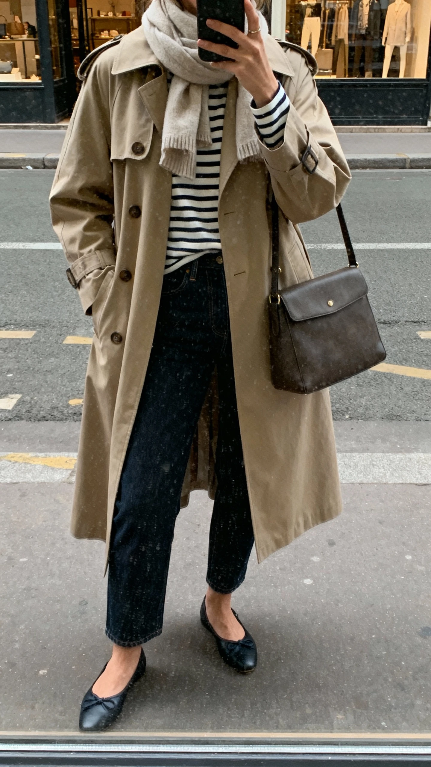 iPhone street-style selfie reflected in a shop window of a woman in a classic trench coat over a Breton stripe top with dark denim and ballet flats, simple scarf and structured bag, face not visible, overcast daylight, natural iPhone photo quality.