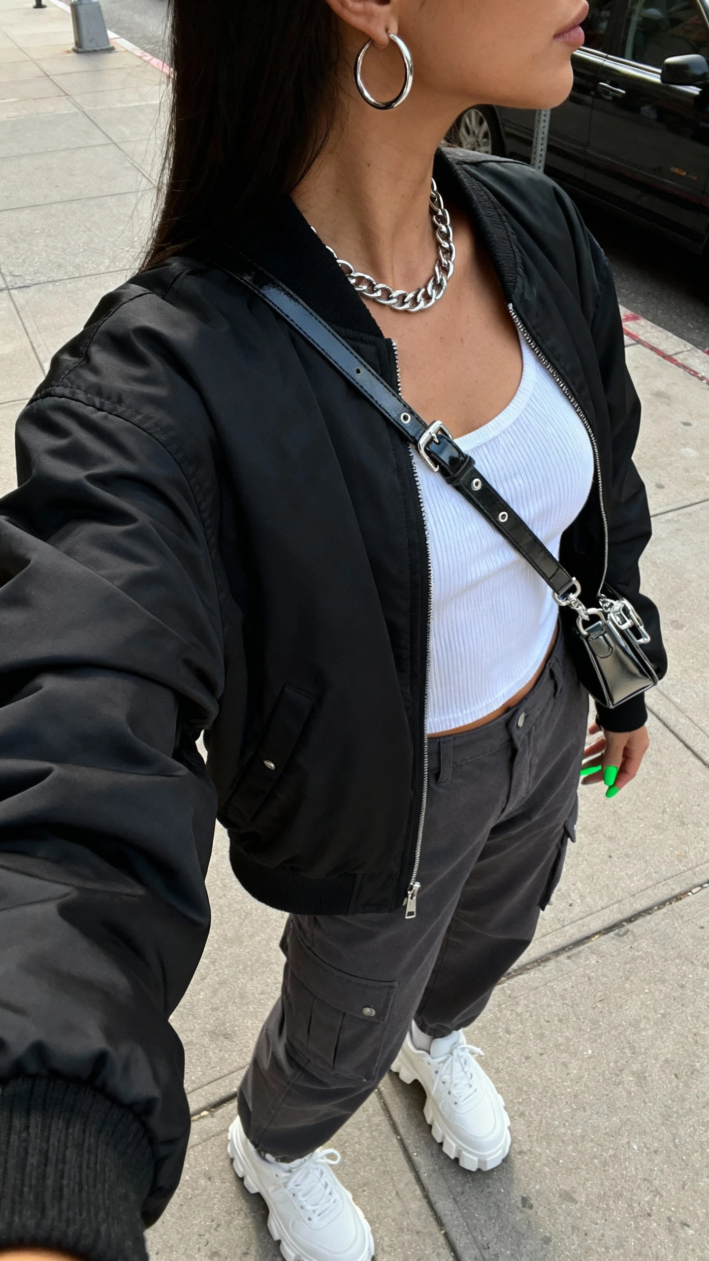 iPhone street-style side-view selfie of a woman wearing city-chic streetwear: oversized black bomber, white ribbed tank, high-waist charcoal cargo pants, white chunky sneakers, silver hoop earrings, layered silver chain, cool green manicure, sleek crossbody with metal hardware; face not visible, natural daylight on a city sidewalk, casual iPhone photo.