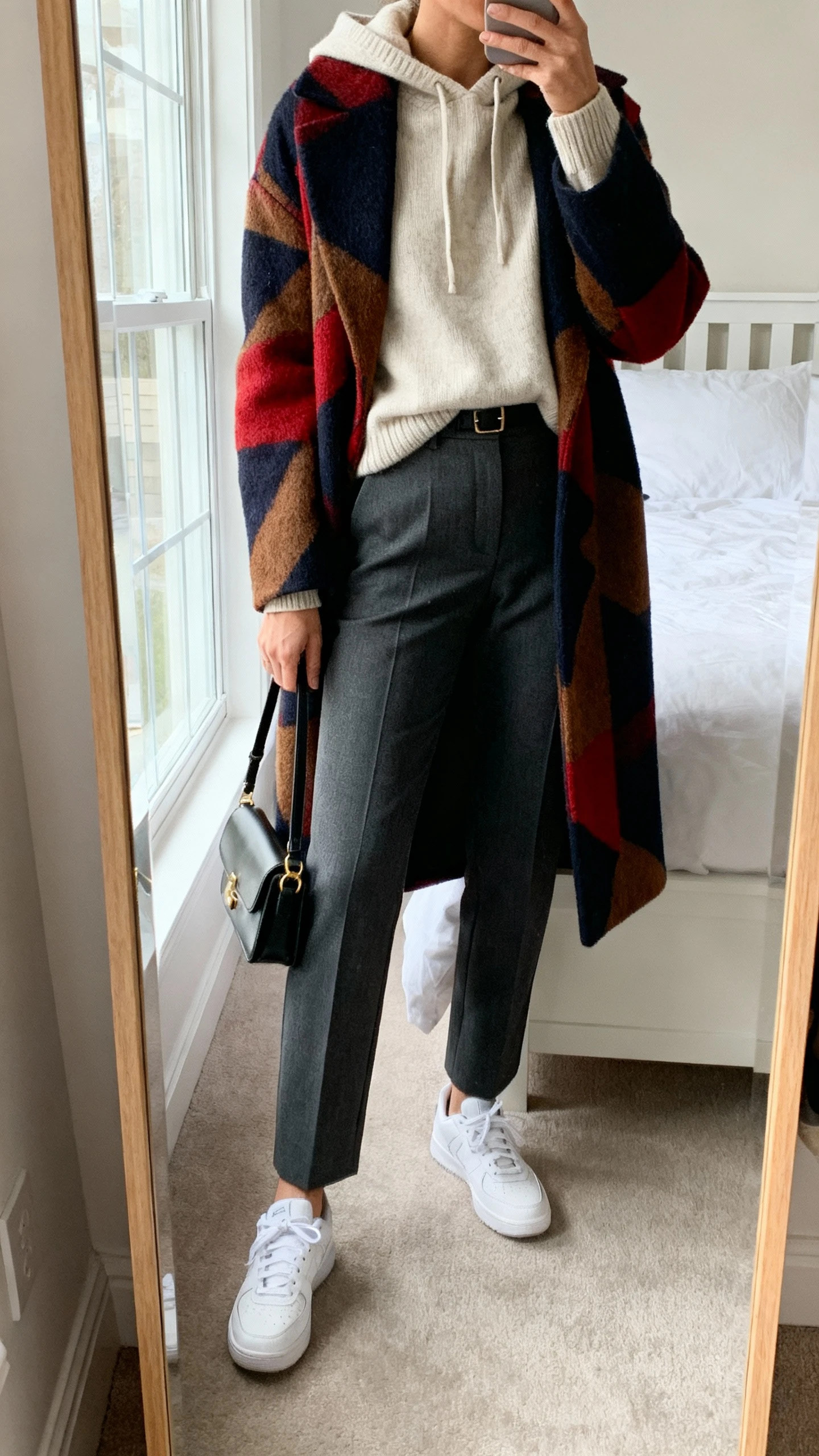iPhone mirror selfie of a woman in an off-duty cashmere hoodie tucked into smart tailored trousers, topped with a bold statement coat and clean white sneakers, small shoulder bag in hand, face not visible, cropped at shoulders, bedroom mirror, natural window light, iPhone photo quality.