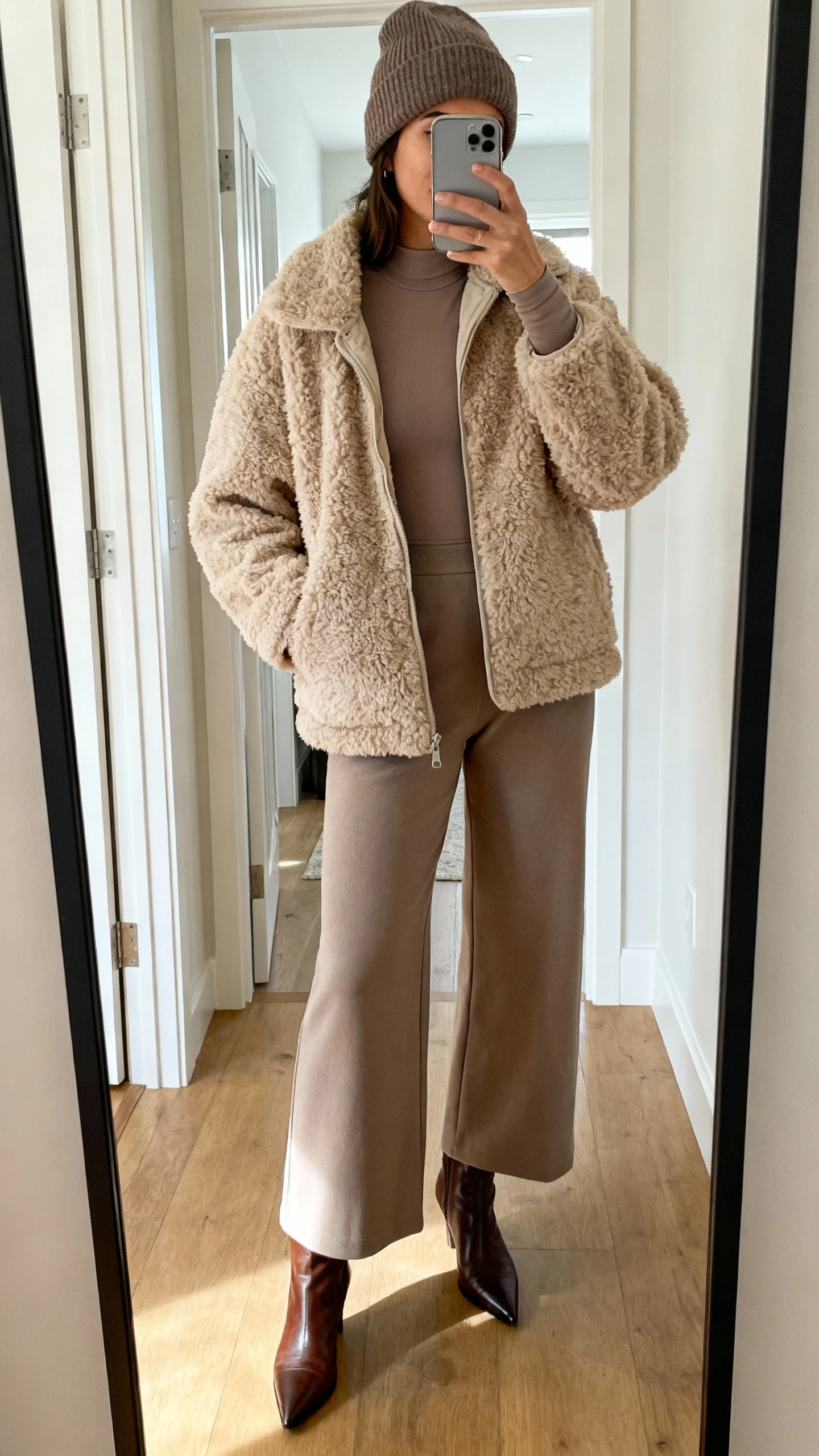 iPhone mirror selfie of a woman wearing a plush teddy shearling jacket over a sleek base layer (fitted long-sleeve top) with wide-leg pants, pointed leather booties, and a beanie, face not visible, cropped at neck, entryway mirror, indoor daylight, iPhone photo quality.