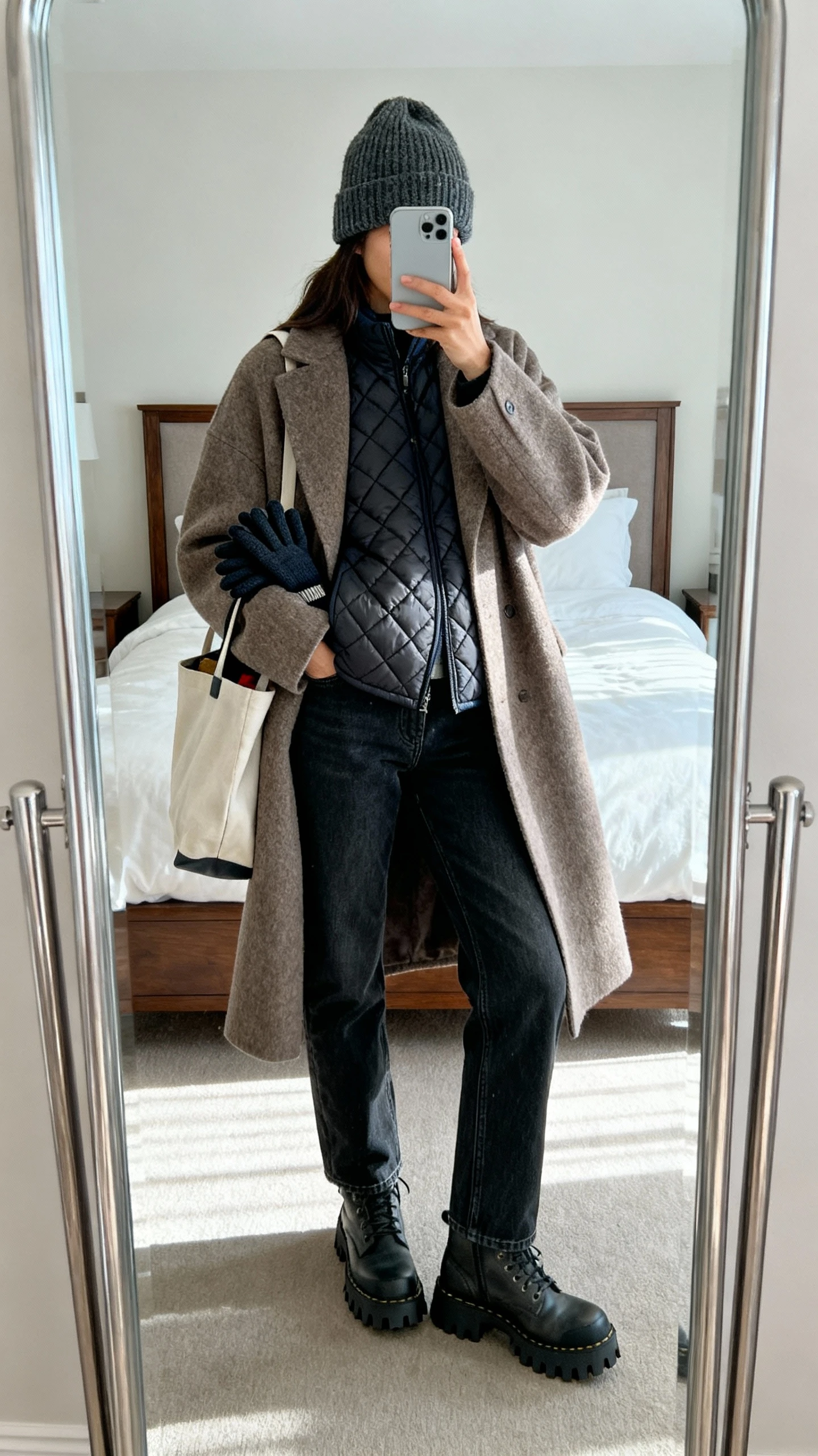 iPhone mirror selfie of a woman wearing a quilted liner jacket layered under an oversized wool coat, straight dark jeans, chunky lug-sole boots, knit beanie, and gloves peeking from a tote, face not visible, cropped at shoulders, bedroom full-length mirror, cool morning light, iPhone photo quality.