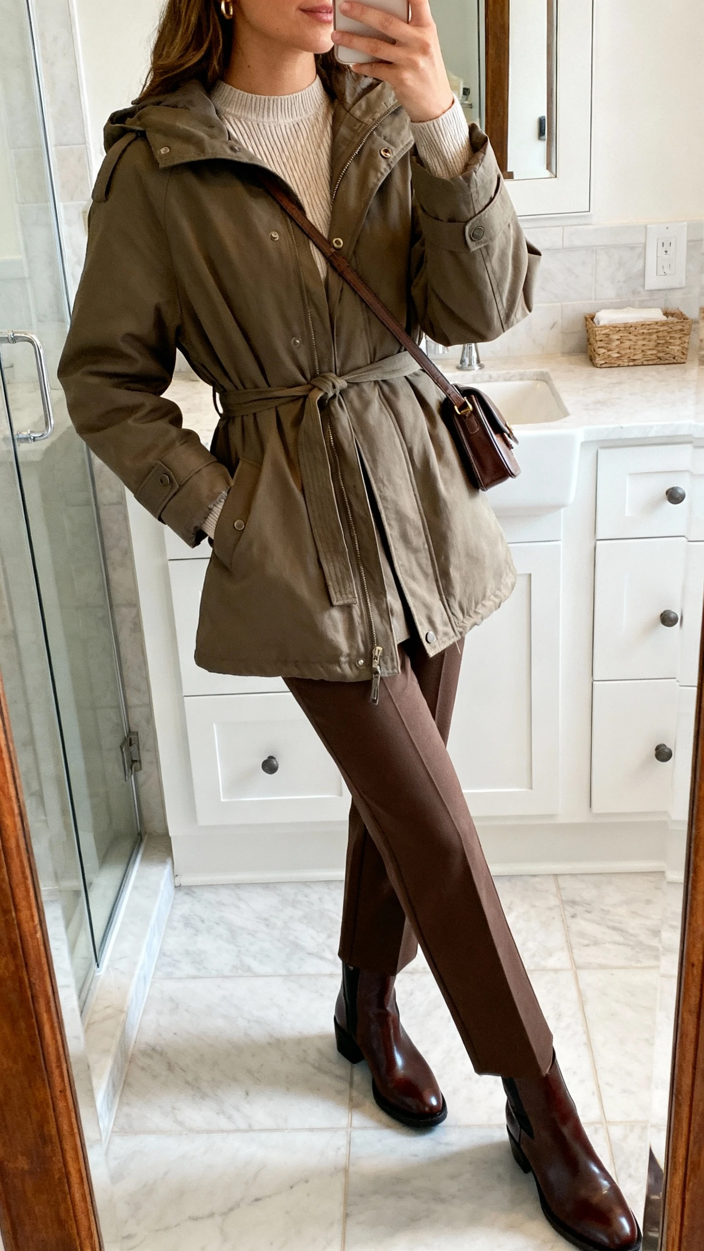 iPhone side-angle mirror selfie of a woman in a belted parka over a fine-gauge crewneck, straight-leg trousers, sleek leather ankle boots, and a refined crossbody, face not visible, bathroom mirror, indoor natural light, iPhone photo quality.