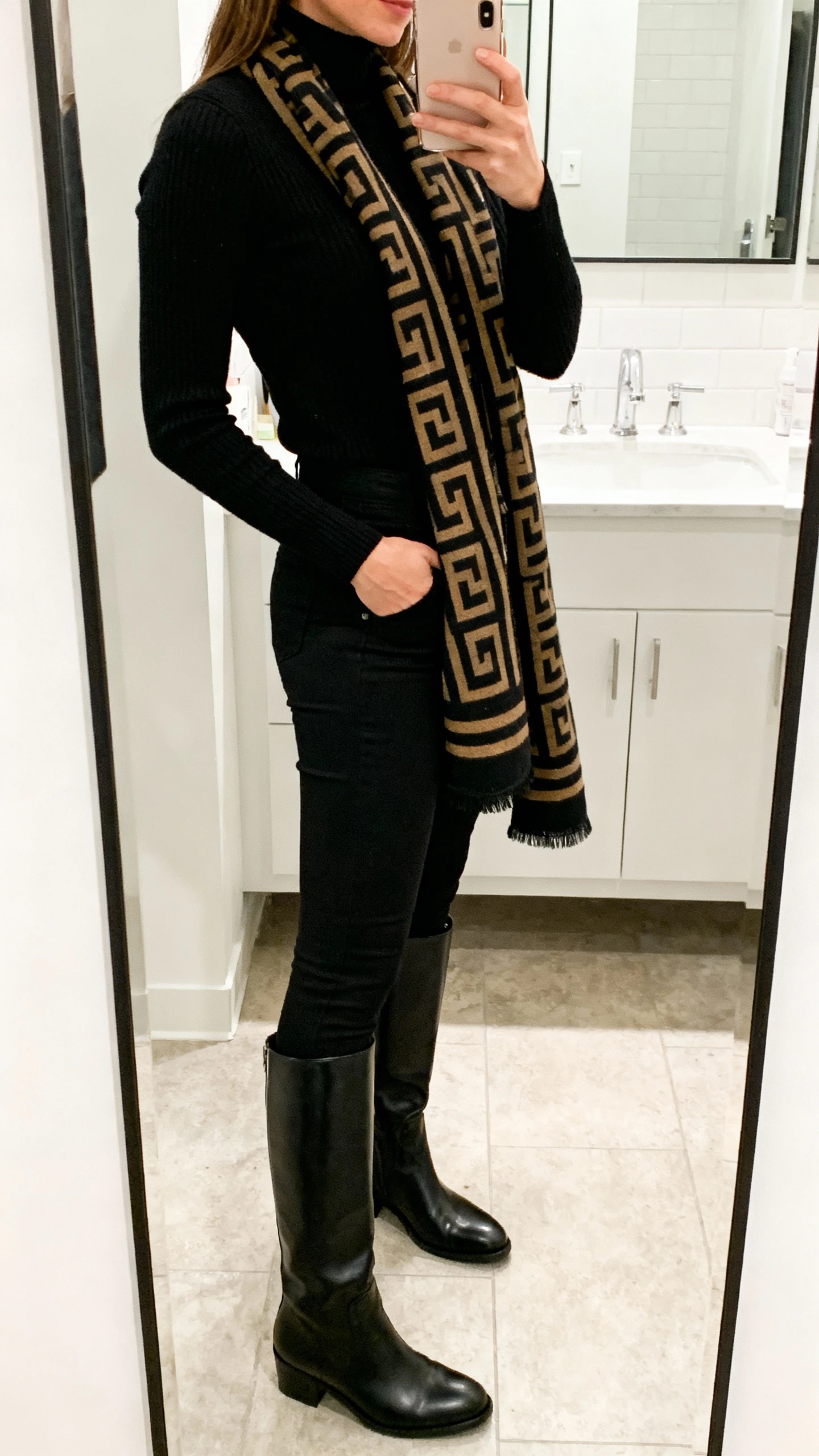 iPhone side-angle mirror selfie of a woman in an all-black base (black turtleneck and black slim trousers) with sleek tall boots and a bold power scarf draped at the neck, face not visible, bathroom mirror, bright indoor lighting, iPhone photo quality.