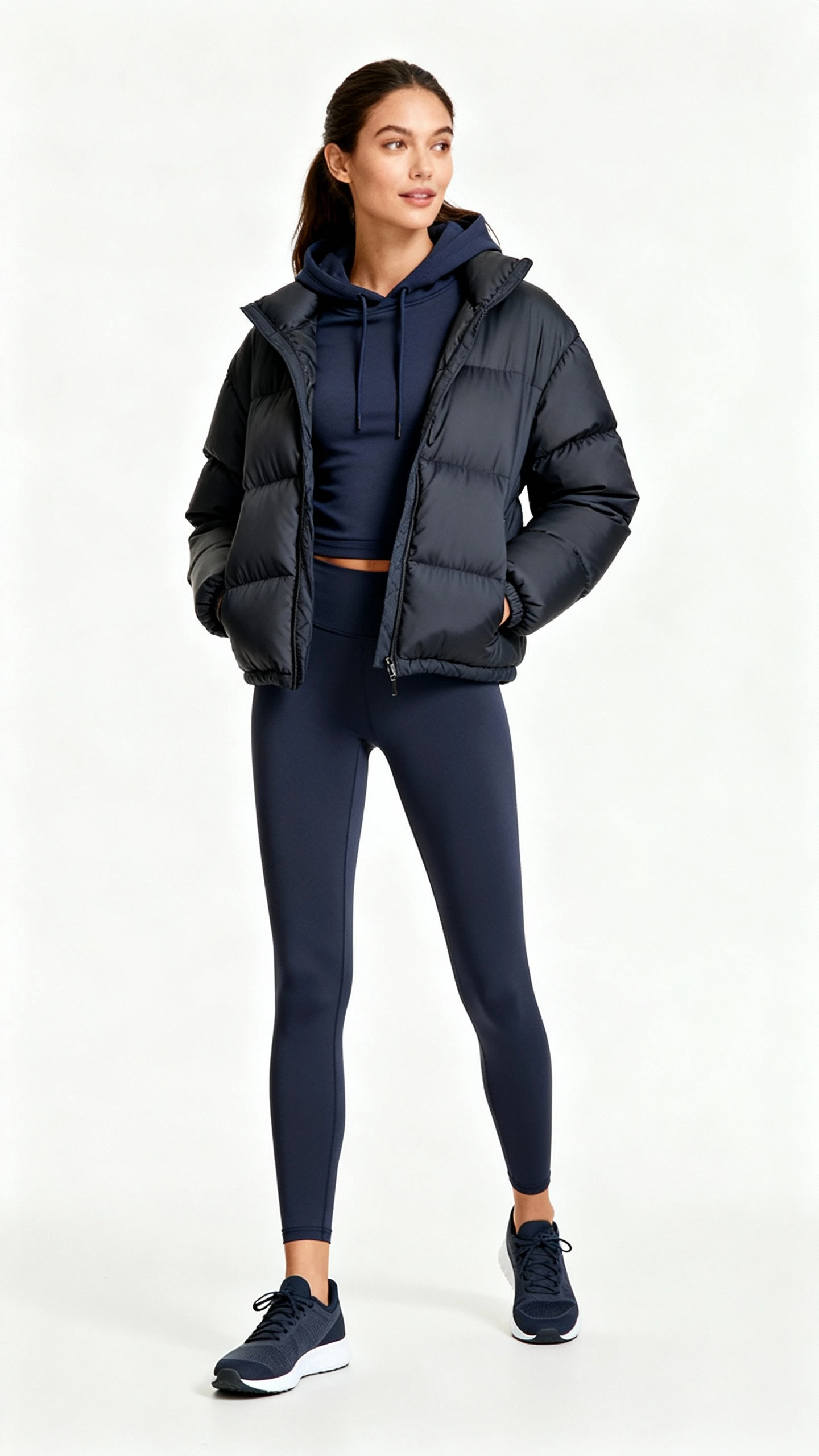 Studio fashion photo of a woman wearing a matte puffer jacket layered over a fitted hoodie and leggings with streamlined sneakers, white background, soft studio lighting, hands in pockets, clean composition, polished athleisure.